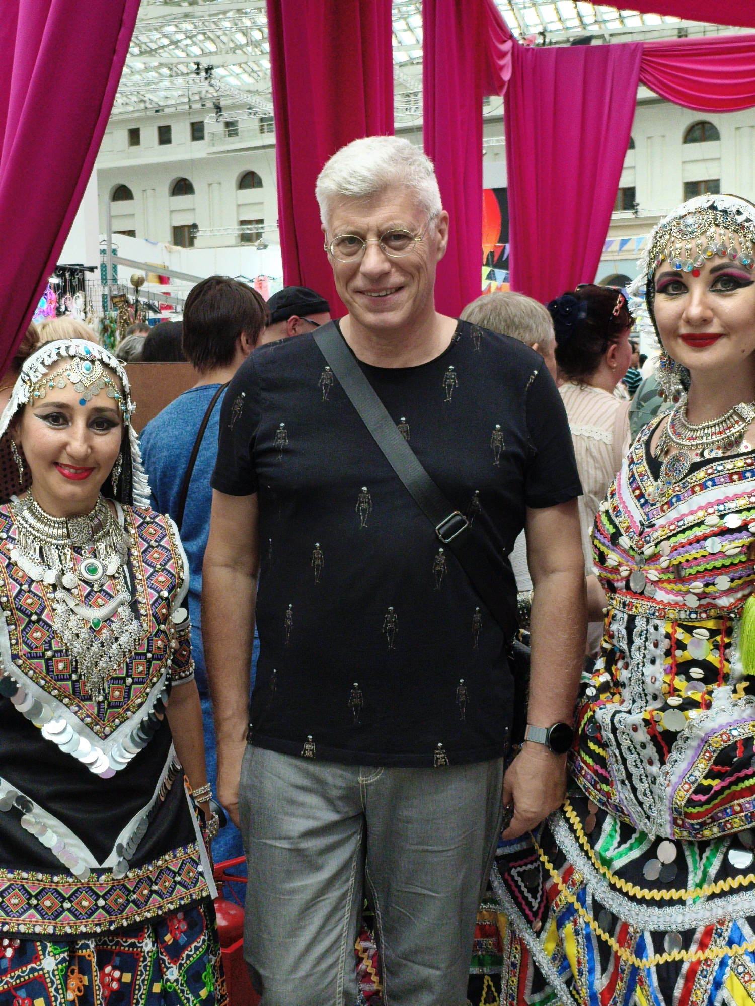 A group of people, including two women in elaborate, traditional costumes and one man in casual attire, are posing for a photograph indoors. The women are adorned with intricate jewelry, including headpieces, necklaces, and bracelets, and wear vibrantly colored, embroidered outfits. The man, wearing glasses and a black t-shirt with a skeleton pattern, stands between them, a crossbody bag slung over his shoulder. The background reveals a large, open space with a high, glass-paneled ceiling, suggesting a public venue like a market hall or exhibition center. The man is wearing a smart watch on his left wrist. The location is identified as Moscow, Russia.