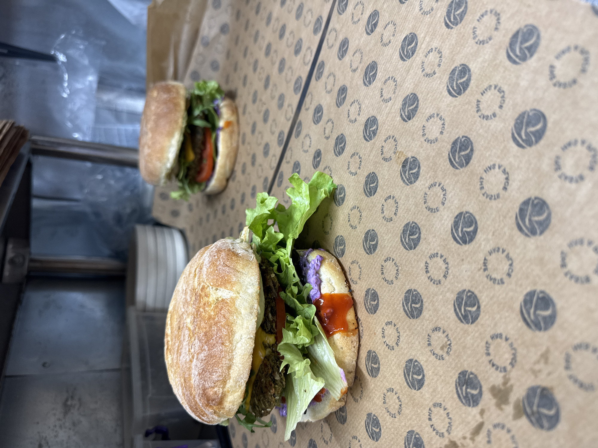 Two sandwiches are displayed on a printed cardboard surface, likely a box or container. The sandwiches are assembled with lettuce, tomato, and what appears to be sauce. The background is blurred, but suggests an indoor setting. The location is Unknown, Unknown.
