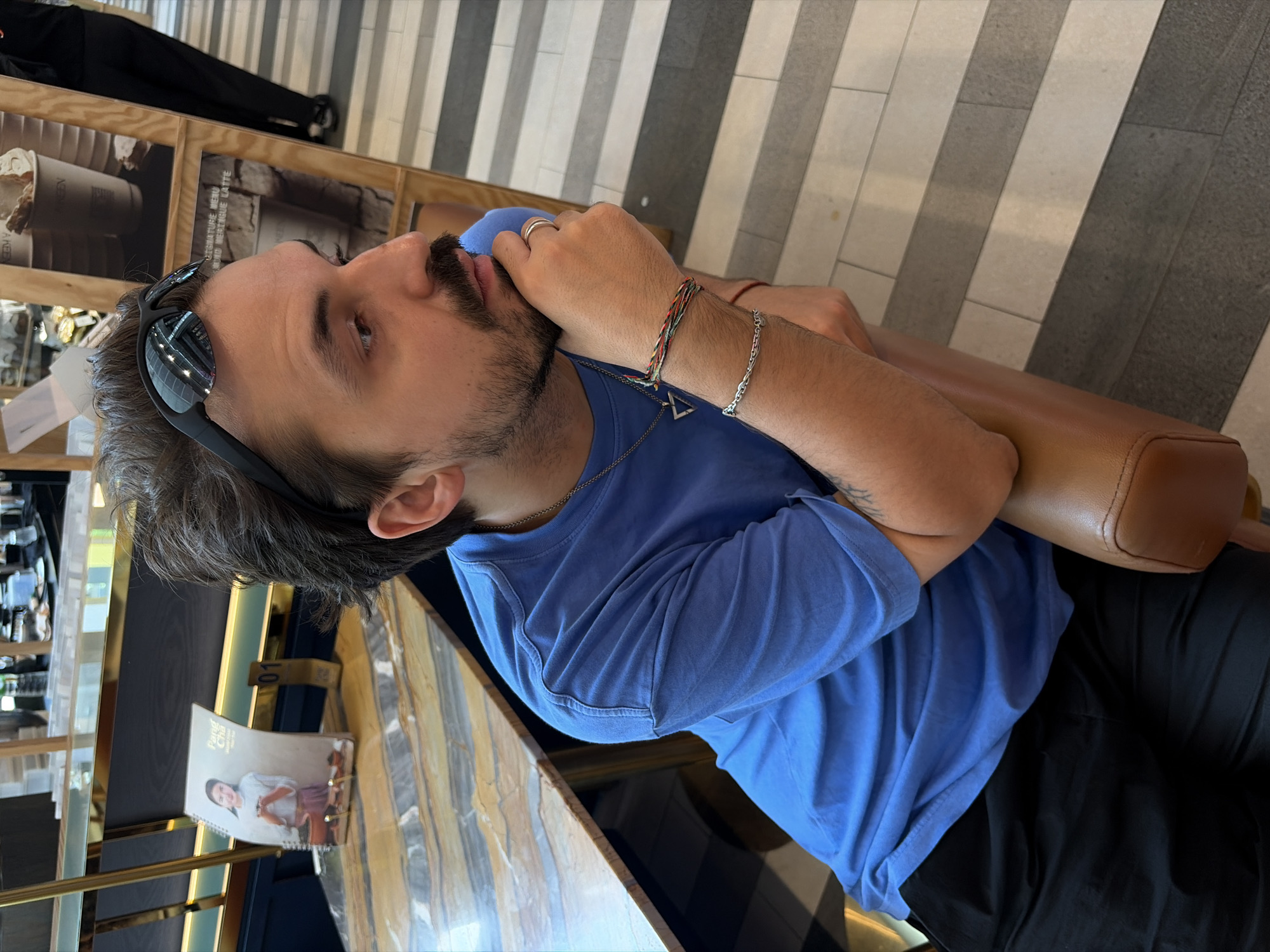 A medium shot captures a man seated inside a building, likely a cafe or restaurant. The man is wearing a blue T-shirt and dark pants, with sunglasses perched on his head. He has a beard and is wearing multiple bracelets on his left wrist. His right hand is near his face, possibly touching his lips or chin. The background features a vertical menu stand with spiral binding, a wooden partition with images or advertisements, and a gray and white striped floor. The table in front of him appears to be marble. The location is inferred to be Bangkok, Thailand.