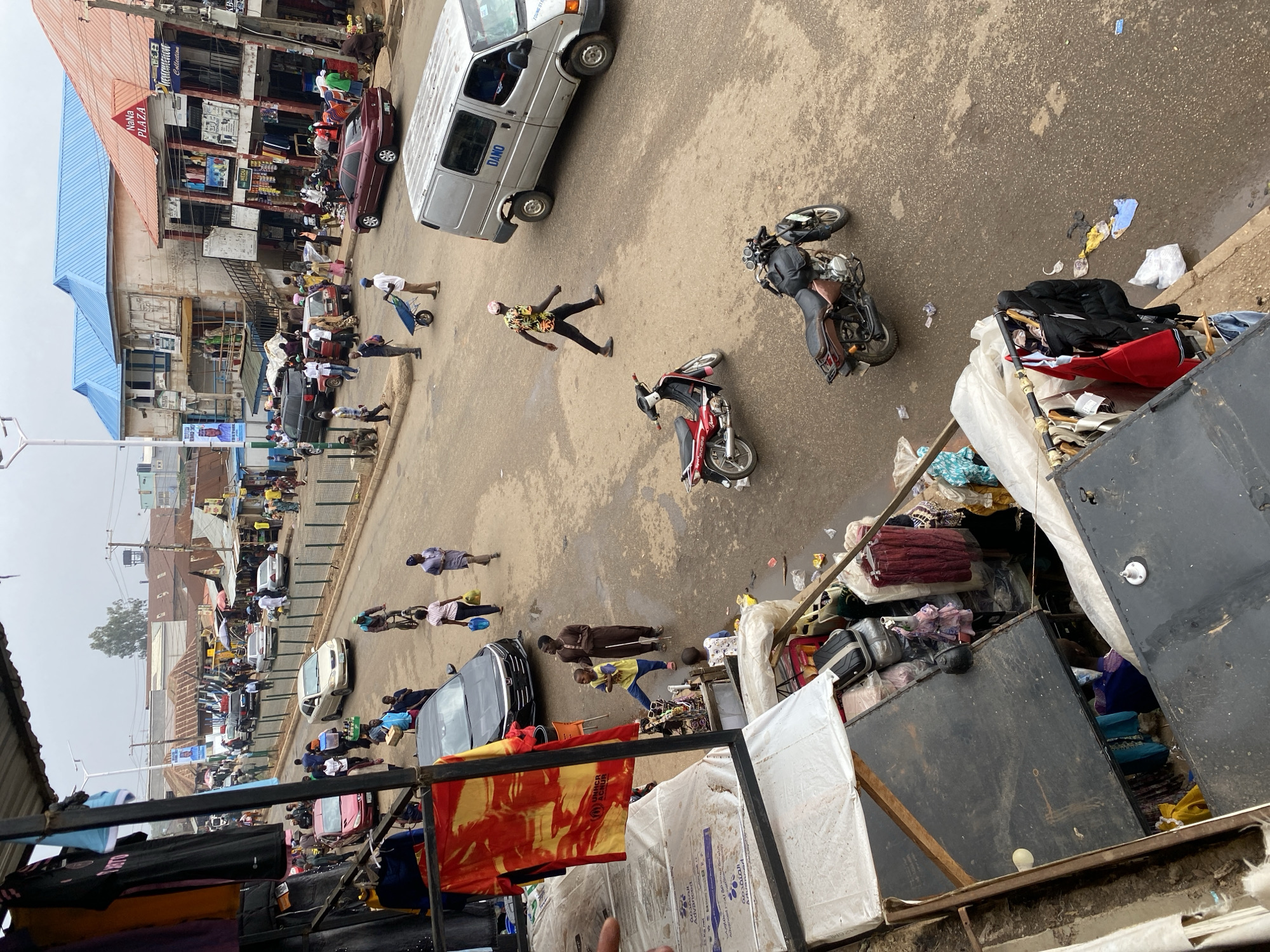 The image depicts a street scene in Bukuru, Nigeria, observed from an elevated perspective. A white van with the inscription "DAMO" is parked diagonally on the street, near a person lying prone on the ground. Two motorcycles are nearby, one on its side. Several individuals are walking or standing along the street and sidewalks, some carrying items or pushing carts. Buildings line the street, displaying various signs and advertisements, including one that reads "NANA PLAZA". In the foreground, there is a chaotic arrangement of objects, including mattresses, furniture, and assorted materials, partially obscuring the view. Clothes are hanging from above, with a visible UNHCR logo. The scene is generally crowded and appears to be a commercial area.