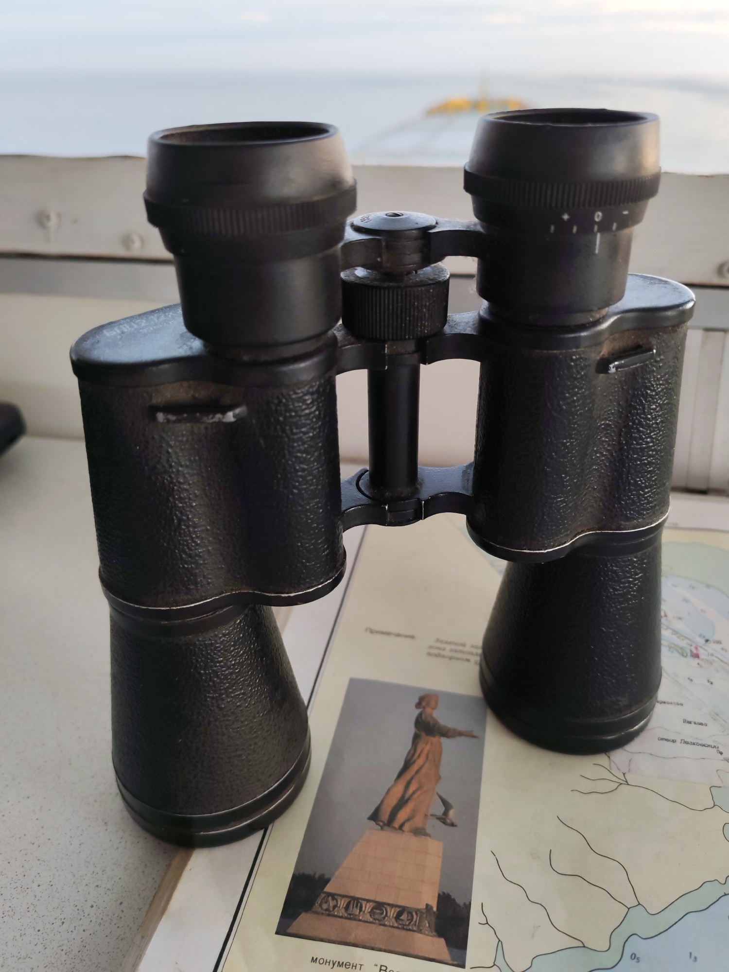 A pair of black binoculars sits atop a nautical chart. A photograph of a statue depicting a woman with an outstretched arm is adjacent to the binoculars. The background shows a blurred, light-colored landscape, possibly a body of water under an overcast sky. The location in the city is unknown, unknown.