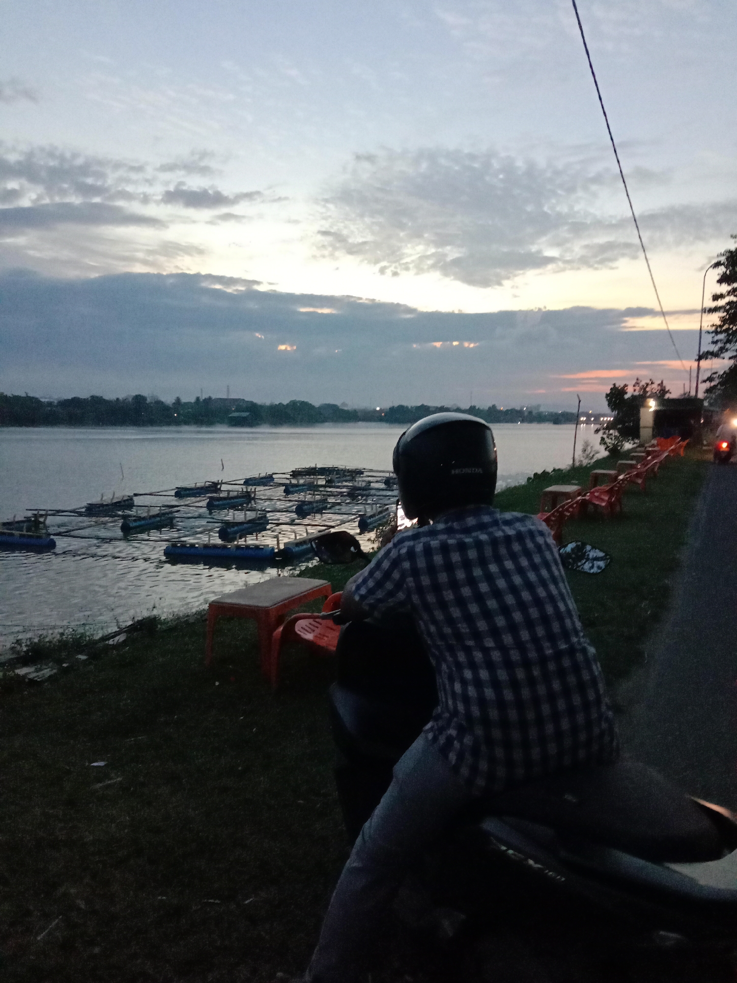 The image shows a person wearing a checkered shirt and a helmet sitting on a motorcycle, facing away from the camera. They are looking out at a body of water with a series of aquaculture cages or rafts on it. The scene appears to be a waterfront area, possibly a river or lake, with vegetation along the bank. In the background, there are trees, buildings, and a sky with clouds that suggest either sunrise or sunset. There are also some orange plastic chairs visible near the water's edge. The setting is likely a public or recreational area in Lhokseumawe, Indonesia, given the presence of aquaculture and the atmospheric lighting often seen during dawn or dusk in tropical regions. The overall mood of the image is calm and reflective.
