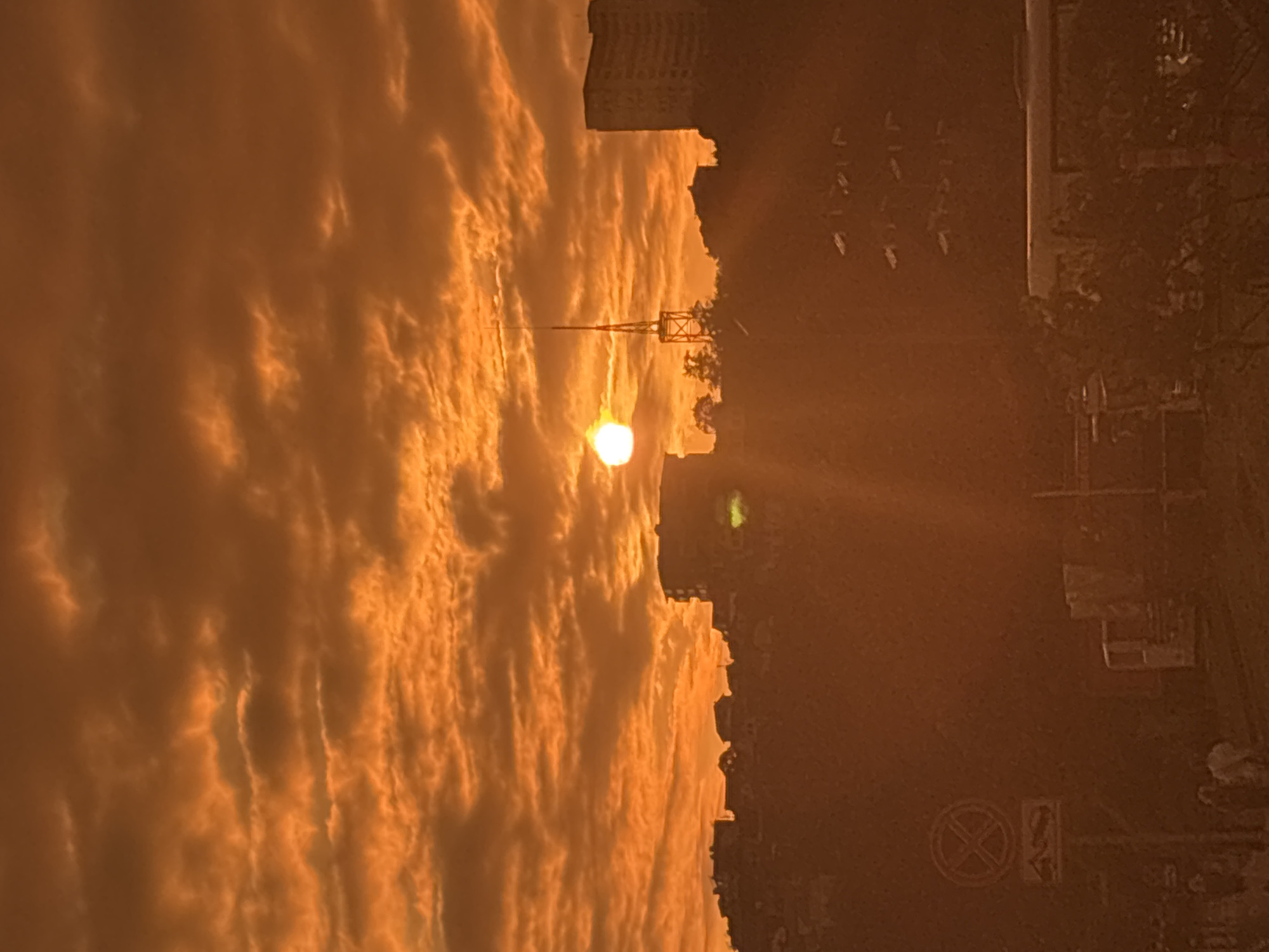 The image depicts a dramatic sunset over the city of Sochi, Russia. The sky is ablaze with vibrant orange and red hues, with textured clouds creating a dynamic backdrop. The sun, a bright white orb, is partially obscured by clouds, casting strong rays of light downwards.
In the foreground, silhouetted against the sky, are the contours of buildings and a distinct radio tower atop a hill. The cityscape appears dense, with various structures ranging from residential buildings to possibly commercial or public facilities. A prominent, tall, slender tower with a lattice-like structure stands out against the horizon, likely a communication or broadcasting tower.
On the left side of the frame, closer to the viewer, a traffic sign is visible, possibly indicating a no-parking zone. The lower part of the image shows hints of street-level activity, with indistinct figures of people walking and what appears to be the side of a bus or large vehicle. The overall impression is one of an urban environment bathed in the intense, warm light of a setting sun, creating a somewhat dramatic and atmospheric scene.