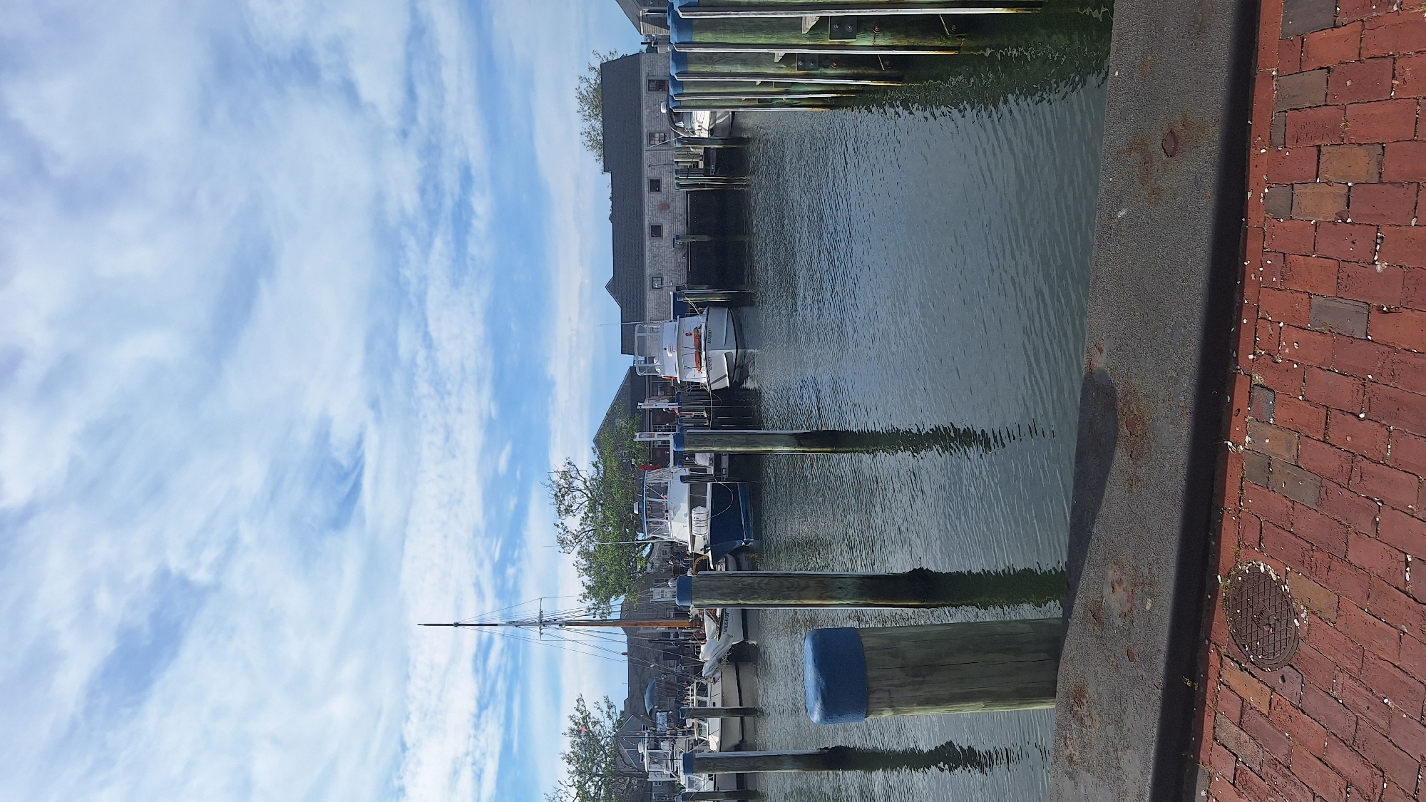 The image depicts a harbor scene in Nantucket, Massachusetts, USA, on a clear day with scattered clouds. In the foreground, a brick walkway lines the edge of a dark gray seawall. Several wooden pilings, some with blue caps, rise from the water, marking the edge of the docks.
Beyond the pilings, several boats are docked. A tall sailboat with a mast stands out among a group of smaller motorboats. These boats are various colors, predominantly white and blue, and appear to be moored securely. In the background, weathered buildings with gray siding and dark roofs are visible, suggesting a commercial or residential area typical of a harbor town. Trees with green foliage are interspersed between the buildings. The water is calm, reflecting the sky and the docked boats. No people are visible in the frame.