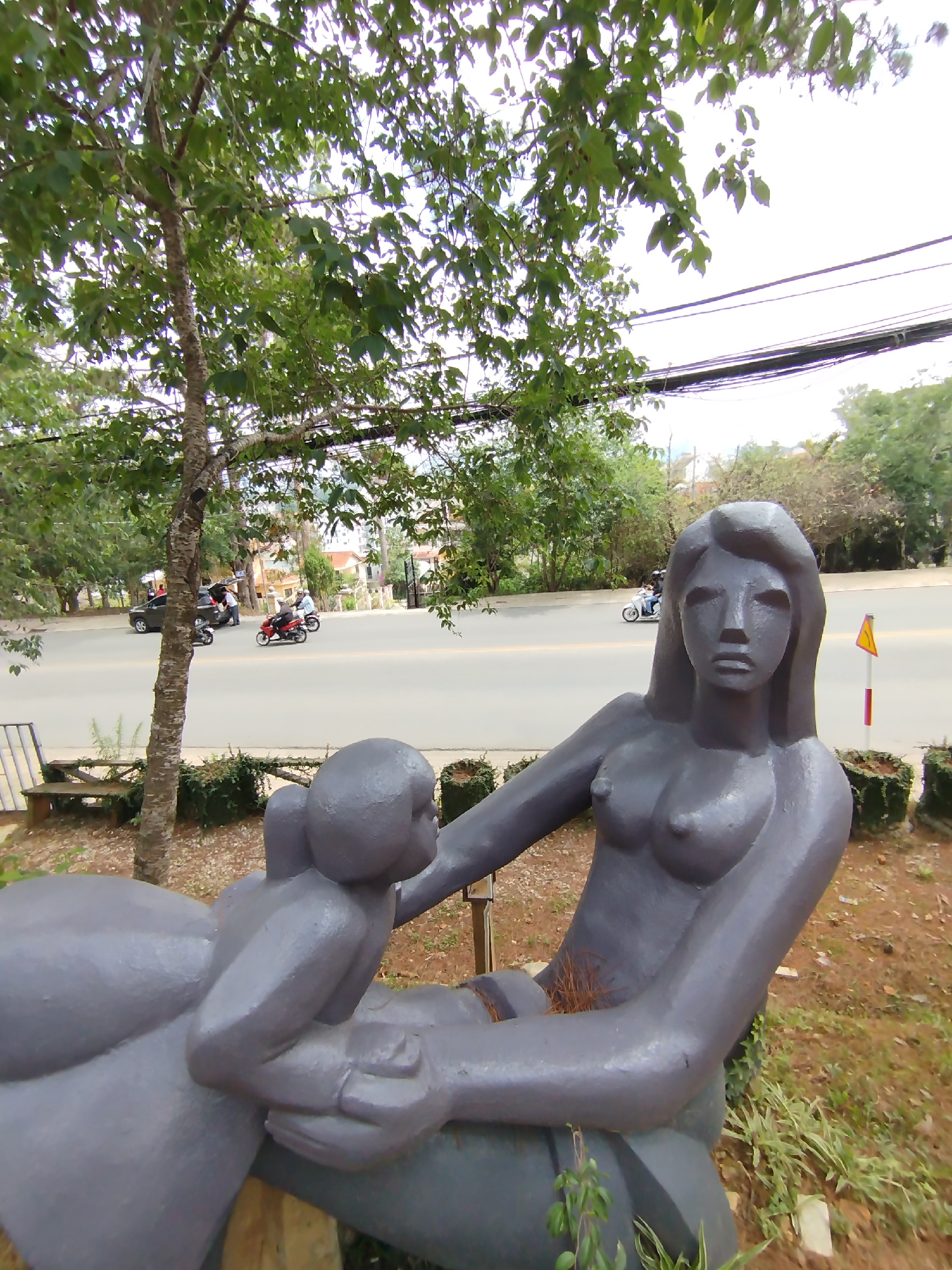 Captured in Da Lat, Vietnam, the image features a grey, nude statue of a woman holding a child, positioned in a garden setting near a roadside. The woman has long hair and a somber expression. A tree stands adjacent to the statue, partially obscuring the view of the road. On the road, motorcycles are in motion. Street-level power lines are visible in the background. The ground is covered with vegetation and reddish-brown earth.