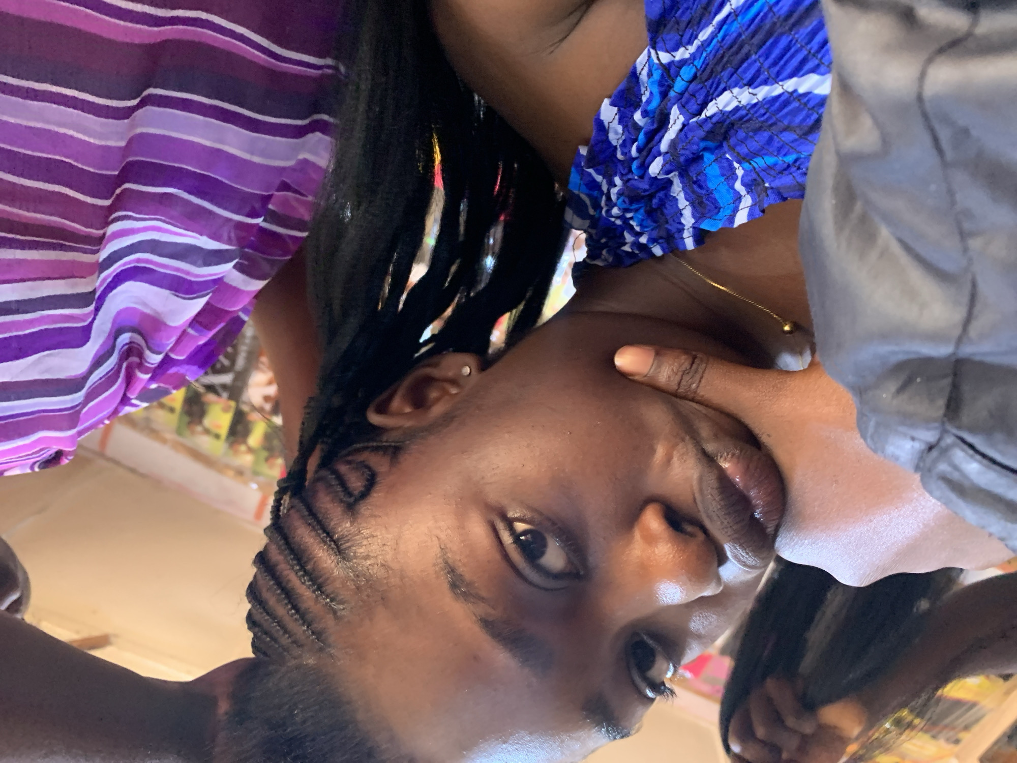 A close-up, slightly angled selfie shows a young Black woman's face and upper body. She is wearing a black jacket on her left side and a blue and white patterned top on her right. Her chin rests on her left hand. Her dark hair is styled in cornrows on the right side of her head, with long black extensions falling down. She is also wearing a thin gold chain with a small ball pendant and has a small stud earring in her right ear. A second person, also Black, is partially visible on the right, wearing a purple and white striped garment. The background is out of focus but appears to be indoors, with hints of colorful posters or displays on a wall. The overall lighting suggests an indoor environment, possibly a salon or a casual setting in New Stadium, Nigeria.