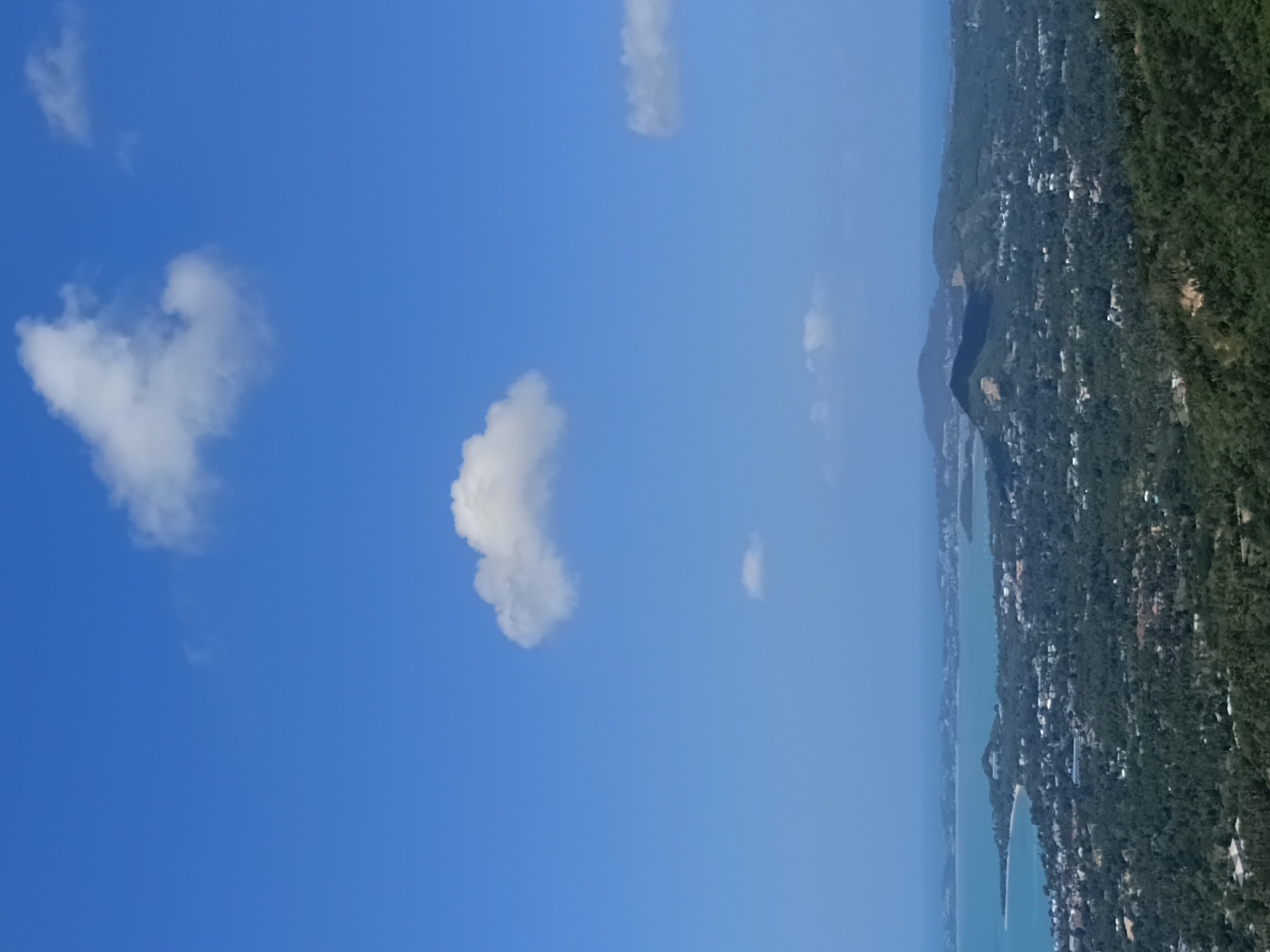 This is an aerial, distant shot of a coastal area in the Ko Samui District of Thailand. The image is dominated by a bright blue sky with several scattered white clouds. Below the sky, a vast expanse of green, undulating hills covers the midground and foreground, dotted with numerous buildings, suggesting a developed area. The coastline stretches across the background, meeting a calm, turquoise body of water. Further out, several islands or peninsulas are visible on the horizon. The overall impression is a scenic, tropical landscape under a clear sky.