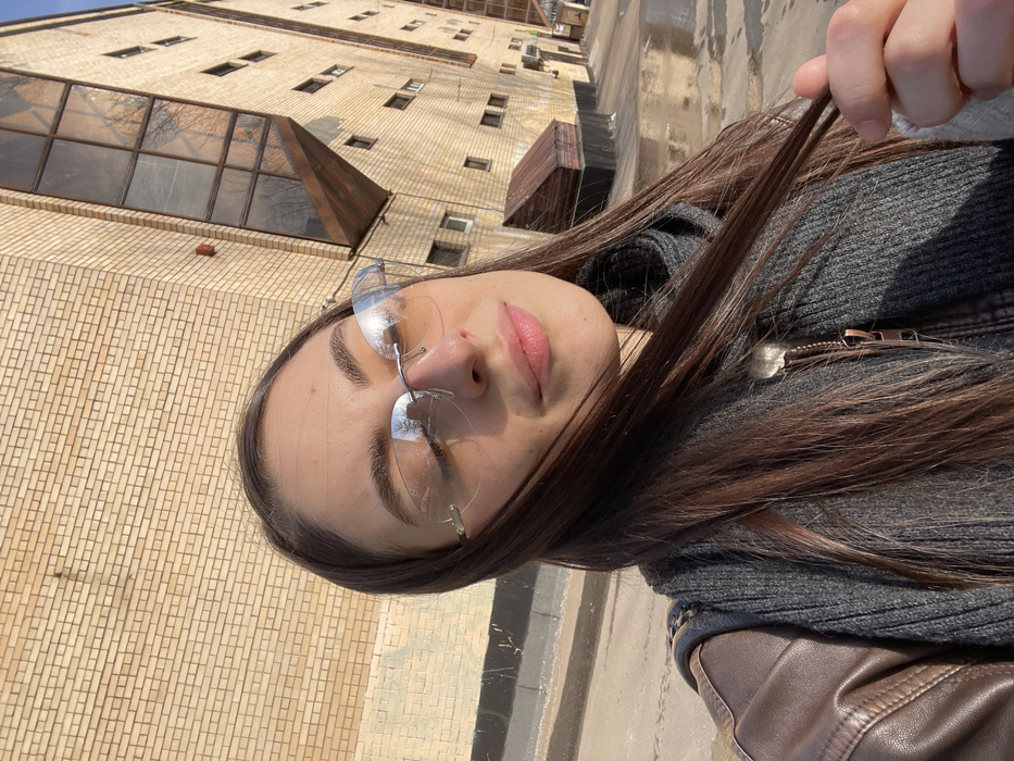 A young woman appears in a medium shot, her eyes partially closed, likely due to direct sunlight. She wears rimless sunglasses with light amber lenses, a brown leather jacket, and a dark grey ribbed knit garment. Her long, dark brown hair is held by her right hand. The background features a large, light brown brick building with a prominent, multi-pane angled window structure on an upper floor, reflecting bare tree branches and the sky. A small, dark brown shed-like structure is at the building's base. The paved ground exhibits wet patches. A clear blue sky occupies the upper right. The location is Bolshaya Semyonovskaya Street, 45, Moscow, Russia.