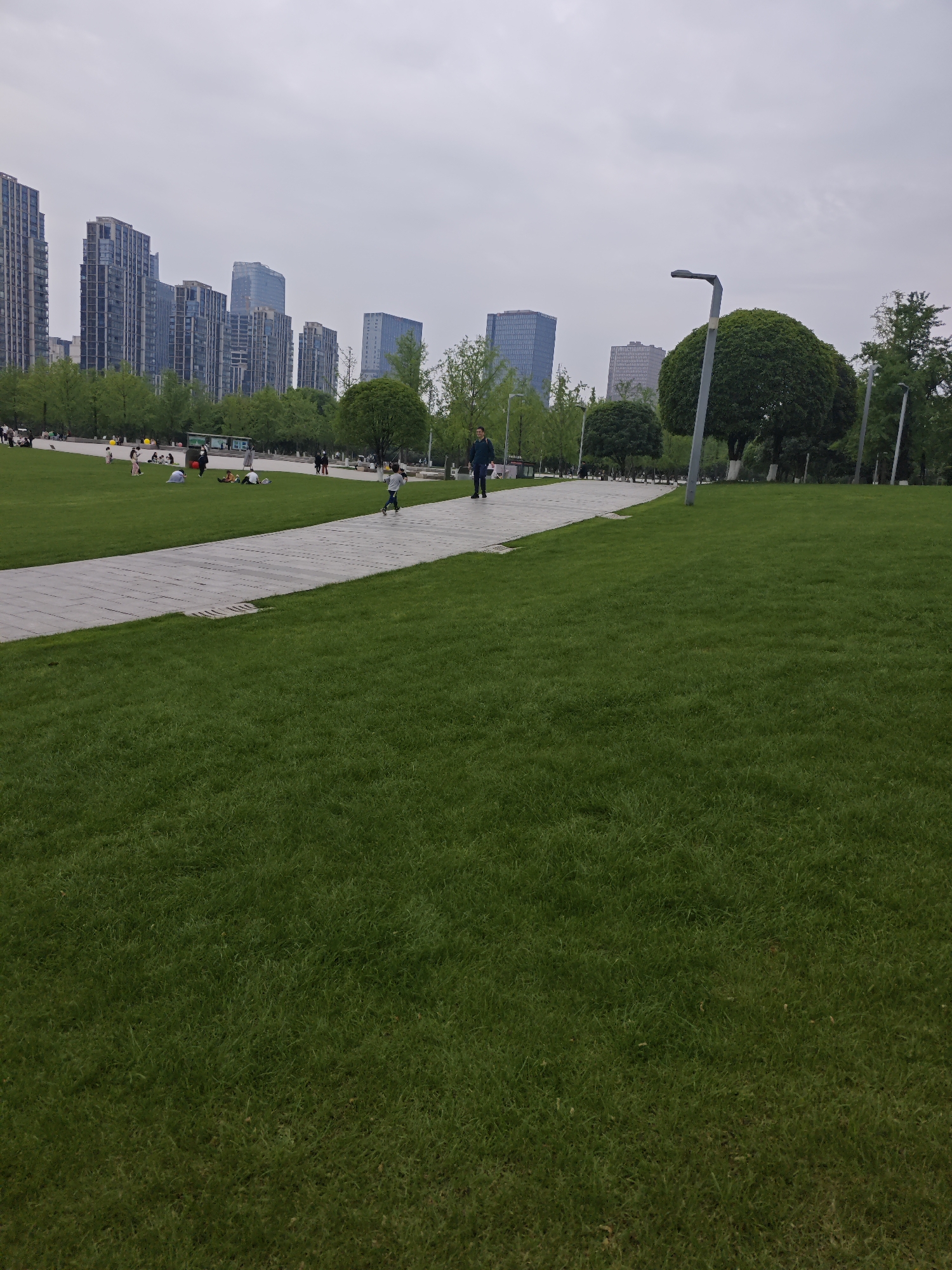 The image depicts a park scene in Liangjiang, China. A wide expanse of lush green lawn dominates the foreground. A paved pathway cuts through the lawn, leading towards the background where several modern high-rise buildings are visible against a cloudy sky. Along the pathway, trees, some with spherical shapes, are planted. A few individuals are walking along the path; a man and a child. Further back, on the lawn closer to the buildings, several people are sitting or lying down. Street lights are positioned along the paved pathway. The overall scene suggests a recreational area within an urban environment.