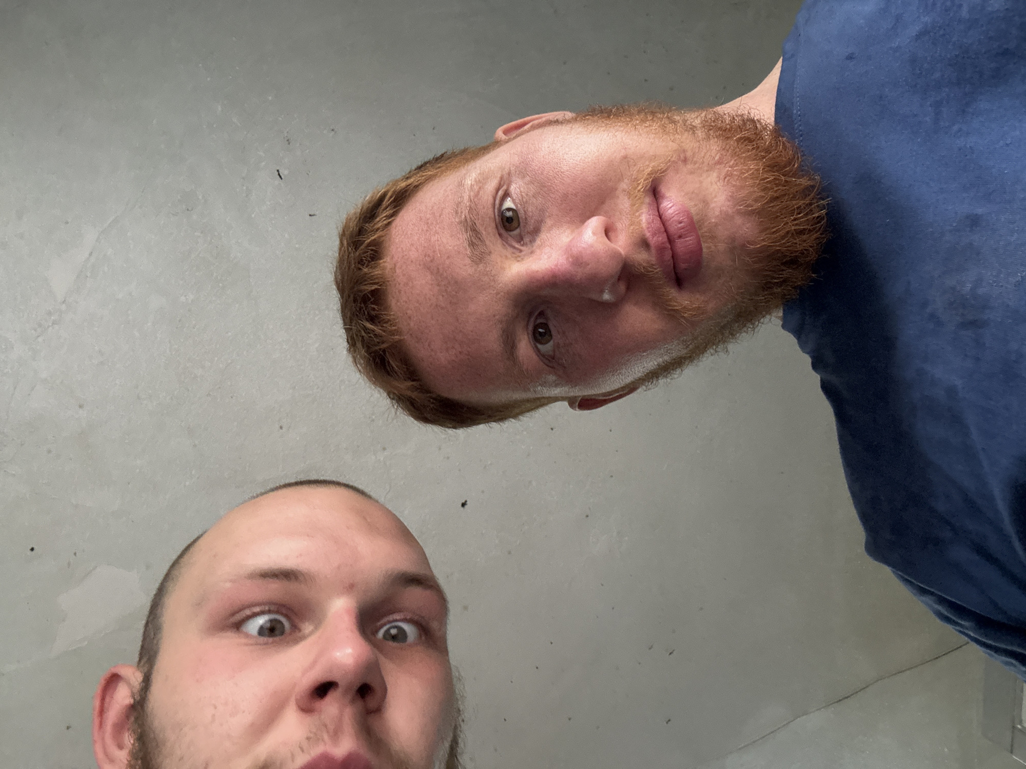 Two men are positioned in a low-angle shot, looking down at the camera. The man on the left, whose head is partially cropped out, has light brown hair and light brown eyes. He is wearing a dark blue t-shirt. The man on the right has red hair and a beard, also wearing a dark blue t-shirt. Both men appear to be indoors, with a concrete-like wall in the background. The lighting is even, suggesting an indoor environment with artificial light.
