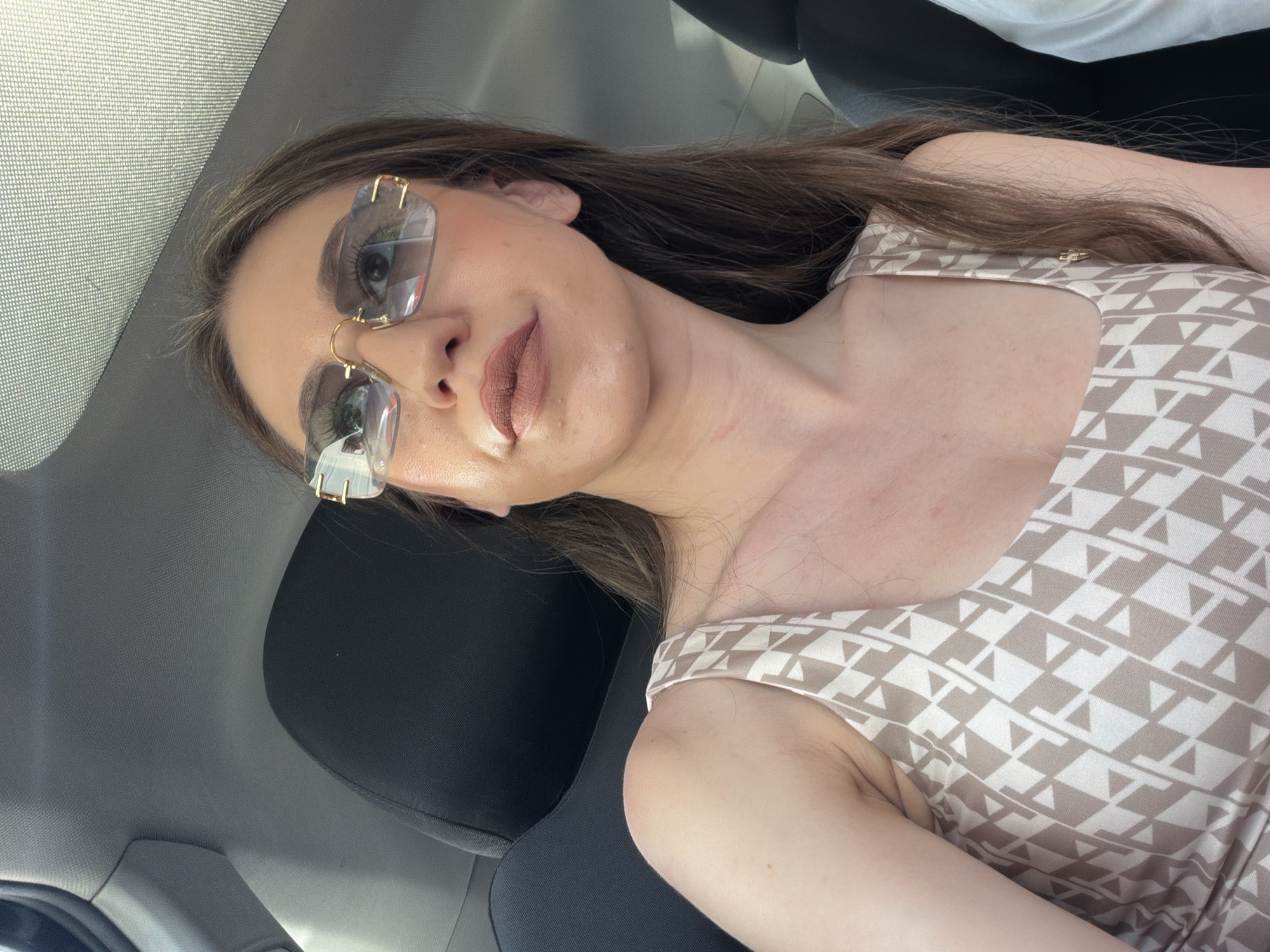 A young woman is seen from the chest up, wearing large, rectangular, rimless sunglasses with a gold frame. She has long, brown hair and is wearing a light-colored tank top with a geometric pattern in beige and white. Her lips are painted with a matte, nude lipstick. The background suggests she is inside a vehicle, with a dark headrest visible behind her and the textured ceiling of a car above. There are no discernible elements in the image that would indicate the location is the city of Sofia, Bulgaria.
