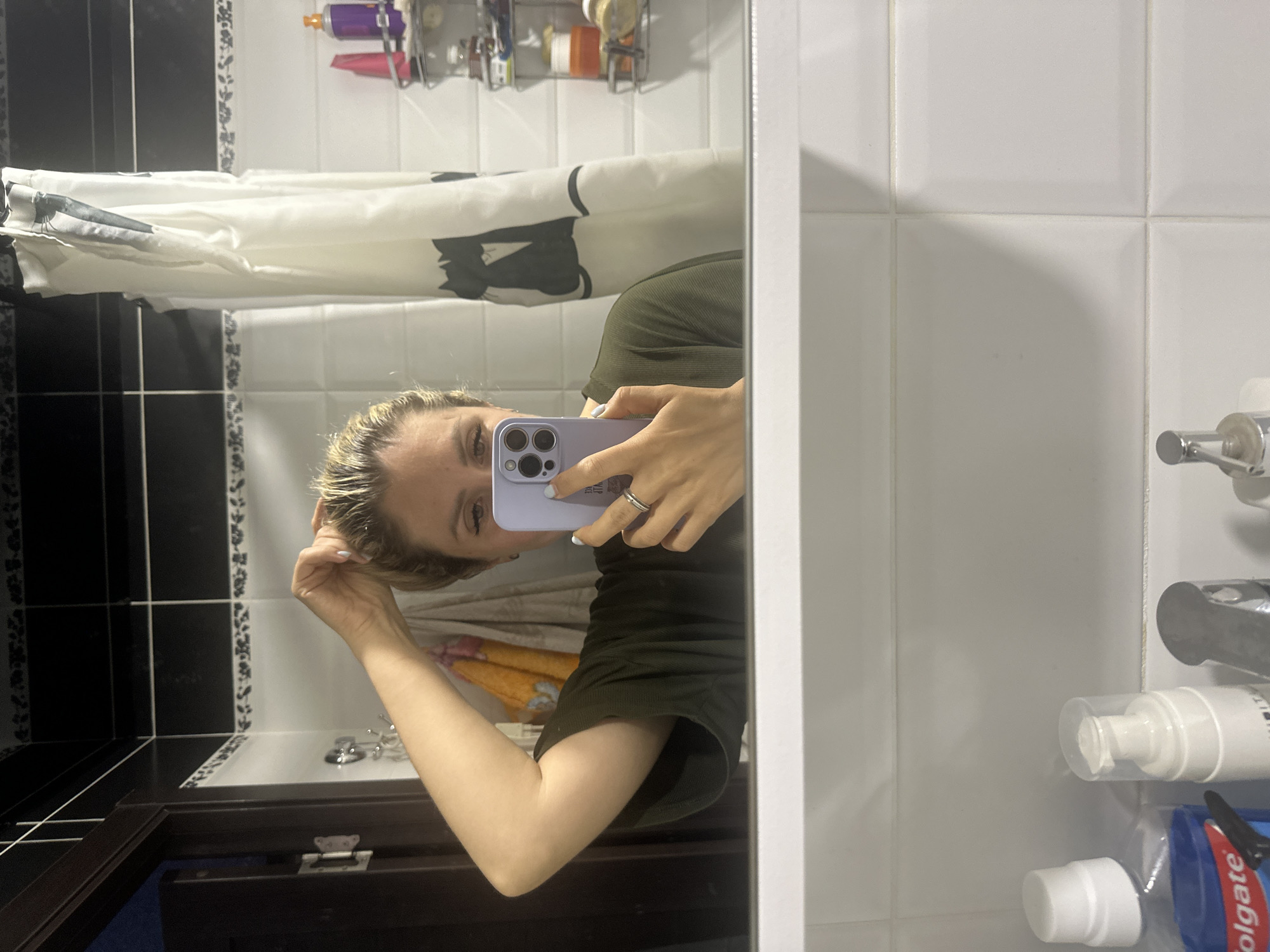 A woman is taking a selfie in a bathroom mirror. She is wearing a dark green t-shirt and has her hair pulled up. Her left hand is holding her phone, which is purple, and her right hand is touching her hair. She is wearing a ring on her left hand. In the background, there is a shower curtain with a black cat design, toiletries on a shelf, and a tiled wall. The reflection in the mirror also shows a sink with various bottles of toiletries. The environment appears to be a typical residential bathroom. The image does not contain any information that would allow for determining its location in Novoaltaysk, Russia.