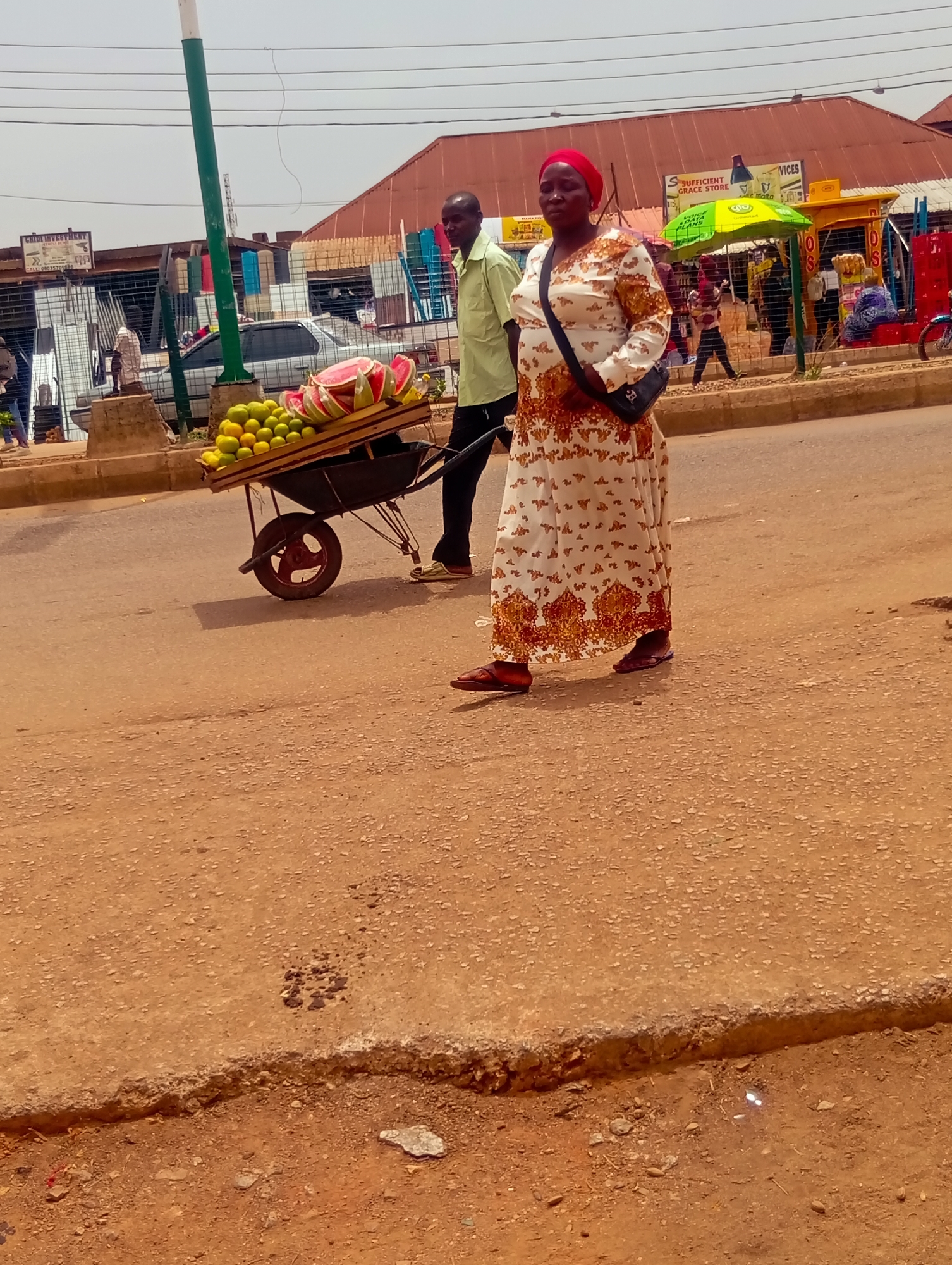 The image shows a street scene in Bukuru, Nigeria. A woman in a long, patterned dress and a red headscarf walks in the foreground, carrying a black shoulder bag. A man is behind her, pulling a black wheelbarrow loaded with oranges and sliced watermelons. In the background, there are buildings, storefronts, vehicles, and other people. A sign that says "Sufficient Grace Store" is visible above the storefronts.