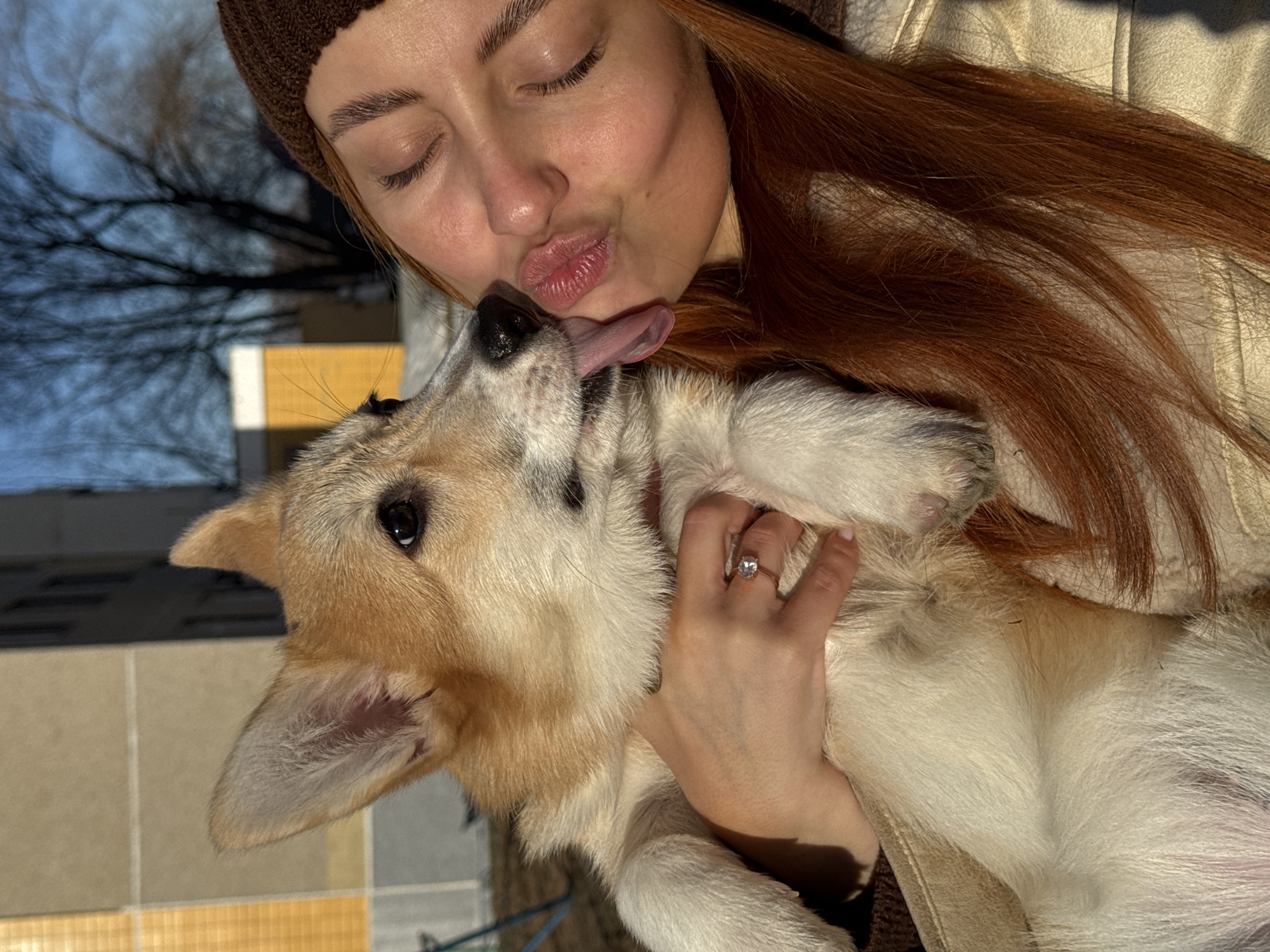 A fair-skinned woman with long red hair and a brown beanie is holding and kissing a tan and white corgi dog. The dog is on its back with its tongue out. The woman is wearing a light-colored jacket and a ring on her left hand. The background shows what appears to be tree branches and a building. This photograph was taken outdoors. Chaikovsky, Russia is not readily discernible in this image.