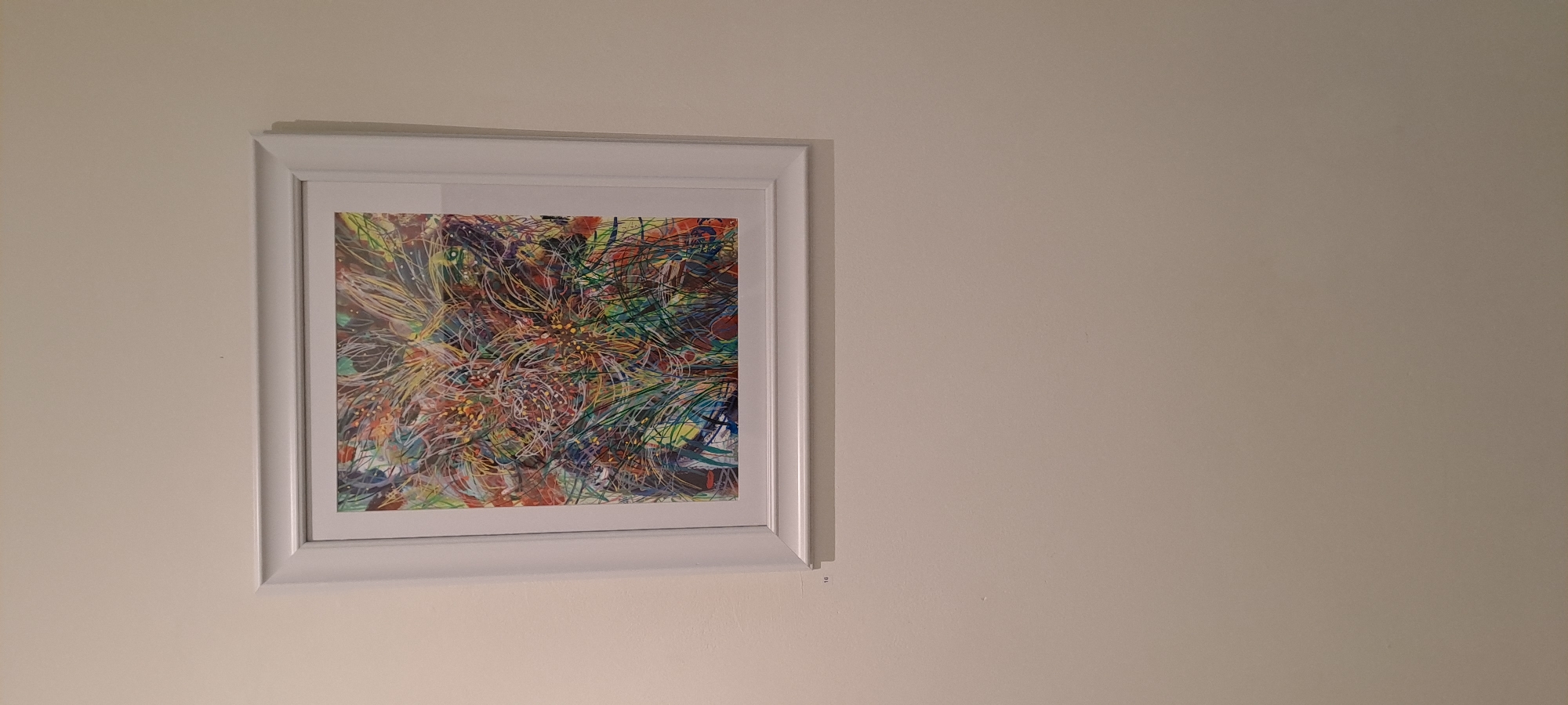 A framed abstract artwork hangs on a plain, light-colored wall. The artwork itself is a vibrant explosion of colors and lines, predominantly featuring shades of green, yellow, red, and blue, with intricate patterns that suggest organic forms or energetic bursts. The frame is a simple, white, rectangular border. Below the artwork, on the wall, a small, discreet number "16" is visible, possibly indicating a catalog or exhibit number. The overall impression is of a piece of art displayed in a gallery or exhibition space. The specific location in Castlebar, Ireland, is not evident from the visual information in the image itself.