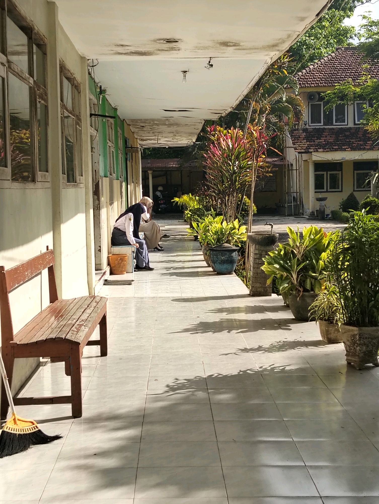 The image shows an outdoor corridor with a tiled floor and a white ceiling that shows signs of water damage and staining. To the left, a light-colored building with several windows is visible, some of which appear to have wooden frames. In the foreground, a wooden bench with a weathered finish is positioned parallel to the building. A broom with a yellow handle and black bristles lies on the floor in front of the bench.
Further down the corridor, two individuals are seated on a bench. One, wearing a light-colored shirt, dark hijab, and trousers, is looking downwards. The other individual, further back and partially obscured, appears to be wearing a long skirt and a dark headscarf. A light brown woven basket is near the seated individuals.
Along the right side of the corridor, several potted plants are arranged. These include large plants with broad green and reddish leaves in dark-colored pots, and smaller plants with thin, grass-like leaves in lighter-colored pots. In the background, a building with a terracotta-tiled roof and yellow walls is visible, with windows and architectural details. Trees and foliage are present around the buildings. The scene is illuminated by natural sunlight, casting shadows from the plants and structures onto the tiled floor. The overall impression is an outdoor walkway at a school or institutional building, likely in a tropical or subtropical climate given the vegetation and light. The specific location in Genteng, Indonesia is inferred by external context not present in the image itself.