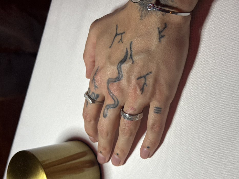 The image displays a close-up, high-angle view of the dorsal side of a human left hand resting on a white tablecloth. The hand exhibits fair to medium skin tone. Multiple black tattoos are present: a prominent stylized snake or wavy line across the back of the hand, various small abstract symbols or runes on the knuckles and dorsal aspect of the fingers, and small black dots on the fingertips. A smooth, silver-colored, hexagonal-shaped ring is worn on the index finger. A silver-colored band ring with a textured, possibly hammered, finish is on the ring finger. A slender, silver-colored cuff bracelet is visible on the wrist. In the upper left quadrant, a cylindrical metallic object, appearing gold or brass in color, likely a lamp base or decorative item, is partially visible. The scene is set indoors, indicative of a dining establishment or similar venue. No discernible elements within the image allow for the identification of a specific location in Yerevan, Armenia.