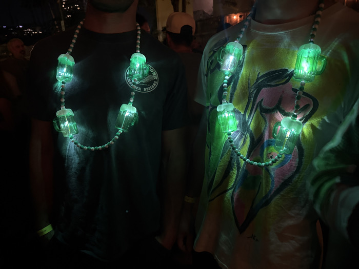 This image depicts two individuals at what appears to be a nighttime social event in Los Angeles, California, United States. The precise location within the city is not discernible from the provided visual information.
In the foreground, two individuals are visible from approximately the chest level upwards. Both are wearing necklaces composed of alternating white and green spherical beads, from which five translucent green, beer-mug-shaped pendants are suspended. Each pendant is internally illuminated with a bright green light.
The person on the left is wearing a dark-colored (likely black or dark grey) short-sleeved t-shirt with a circular light-colored logo or graphic design centered on the chest. A neon green wristband is visible on their left wrist.
The person on the right is wearing a light-colored t-shirt with a multi-colored, abstract graphic design across the front, possibly depicting stylized human figures or shapes. A neon yellow wristband is visible on their left wrist.
Both individuals' faces are cropped out of the frame. The background is dark and blurred, revealing several indistinct figures, indicating a crowd, and scattered ambient lights, consistent with an outdoor or semi-outdoor venue at night.
