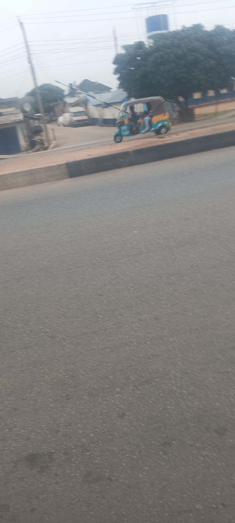 This is an eye-level, outdoor shot capturing a street scene, likely taken from a moving vehicle given the noticeable motion blur across the image. The lighting is soft and diffused, indicating an overcast or hazy daytime.
The lower half of the image is dominated by a grey asphalt road. A dark grey and brown curb separates the road from a paved area running parallel to it on the right side. On this paved area, in the midground, a light blue and yellow three-wheeled auto-rickshaw (commonly known as a Keke Napep in Nigeria) is visible. It appears to be carrying at least one person, though details are blurred. Further along the paved area, a white vehicle is partially seen.
The background consists of various buildings, some featuring blue roofs or blue accents on their structures. A large, leafy green tree stands prominently behind the auto-rickshaw, and a blue cylindrical structure, possibly a water tank, is partially visible behind the tree. Multiple utility poles and overhead power lines crisscross the upper portion of the frame, characteristic of an urban or semi-urban landscape. The scene conveys a sense of daily life in a bustling roadside environment. No discernible text is visible anywhere in the image.