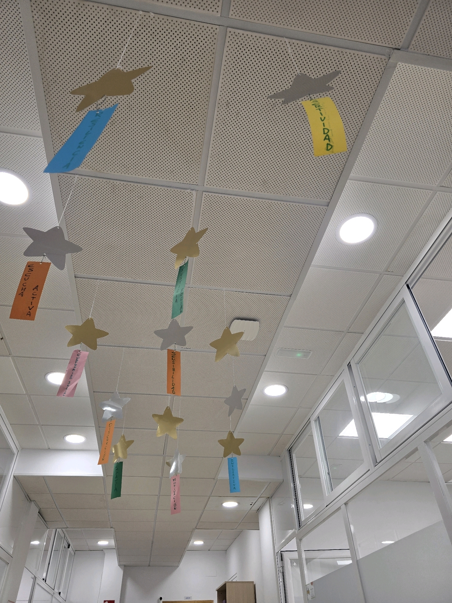 The image shows a ceiling in what appears to be an indoor public space, possibly a school or office building, in Jaén, Spain. The ceiling is made of white, perforated acoustic tiles. Several decorations are suspended from the ceiling by thin white strings. These decorations consist of cut-out star shapes in various colors (gold, silver, tan, and grey) and rectangular paper strips with handwritten Spanish text. Some of the visible text includes "FELICIDAD", "ESCUCHA ACTIVA", "INICIATIVA", "MOTIVACION", and "DESCUBRIMIENTO". There are also circular fluorescent light fixtures embedded in the ceiling. The environment appears to be a corridor or a room with large glass-partitioned walls visible on the right side of the image. There are no people visible in the frame.