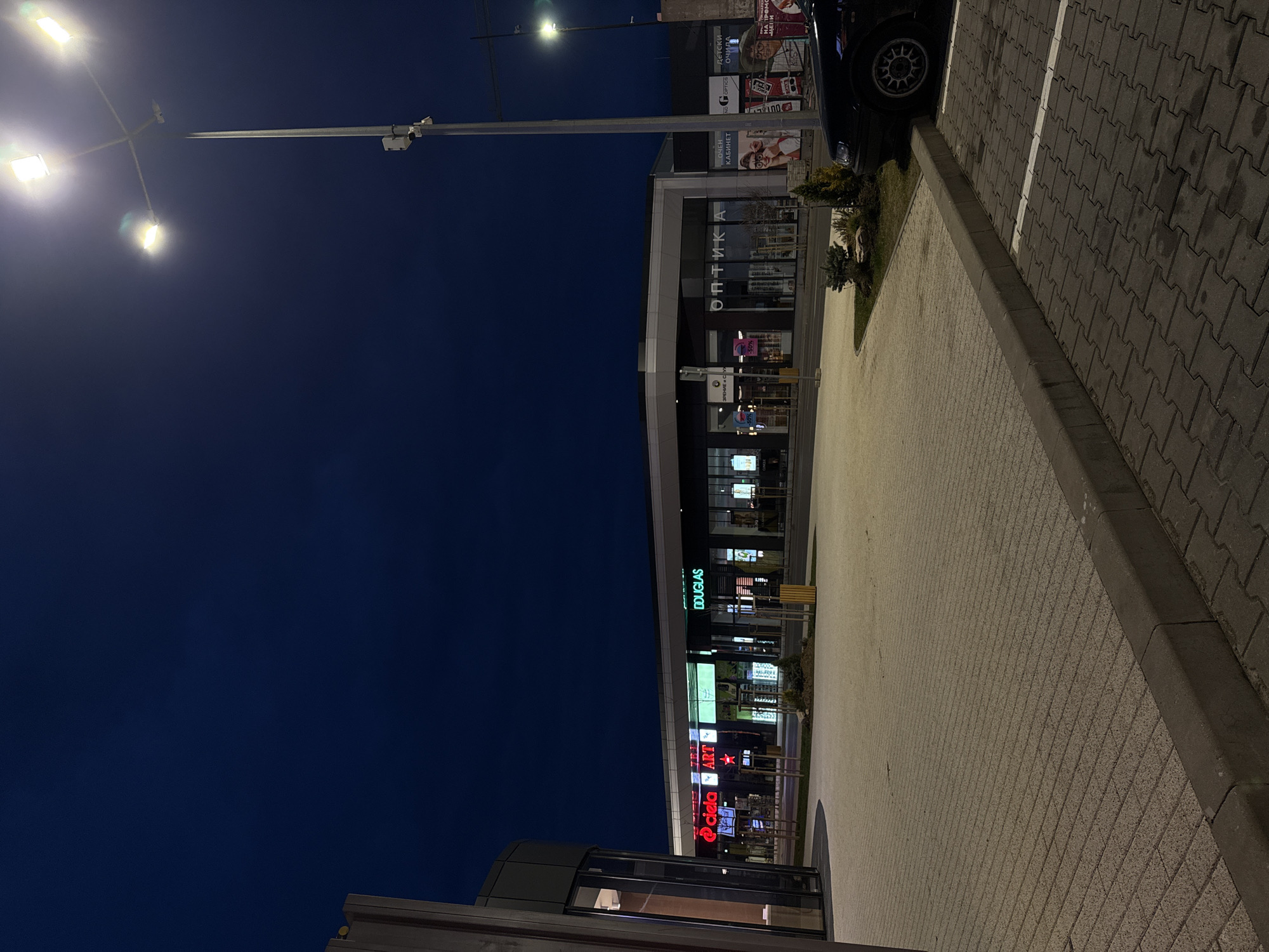 The image shows a street scene at night in Sofia, Bulgaria. A street light illuminates a row of shops and businesses. Shop signs such as "Douglas", "@ Ciela", "Art" are visible. A car is parked along the sidewalk. The street is paved with stone blocks. The sky is dark blue. The overall impression is of a commercial area at dusk.