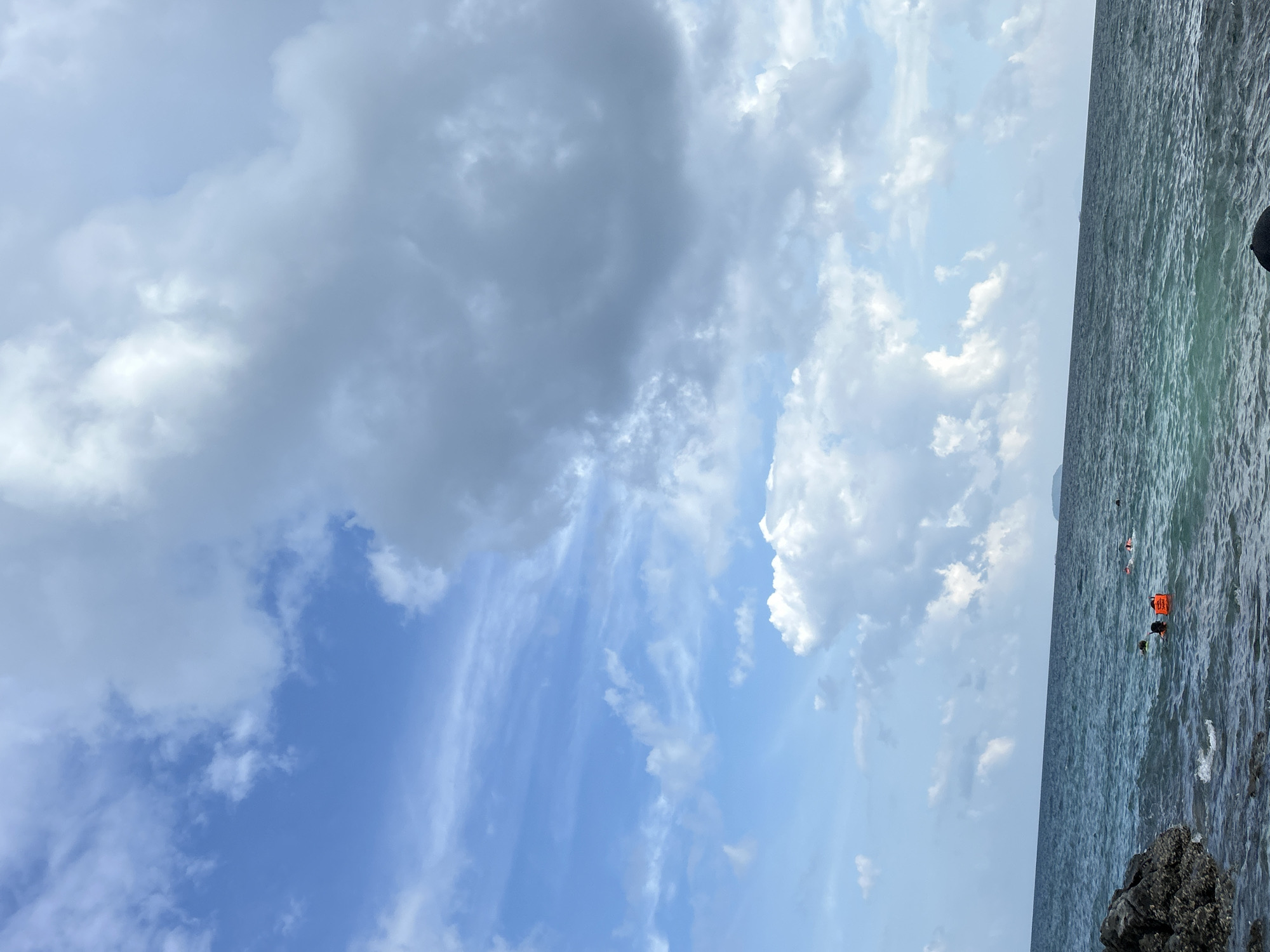 The image shows a partly cloudy sky over the ocean near Phru Nai, Thailand. The sea is a deep blue with moderate waves. In the foreground, there are dark, rocky formations on the left side of the frame, partially submerged in the water. Several individuals are visible in the water, engaged in snorkeling or swimming. One person is wearing an orange life vest. In the distance, the horizon is marked by a faint outline of land. The sky is a mix of bright blue patches and large, gray clouds, with some lighter, white clouds scattered throughout. The lighting suggests it is daytime.
