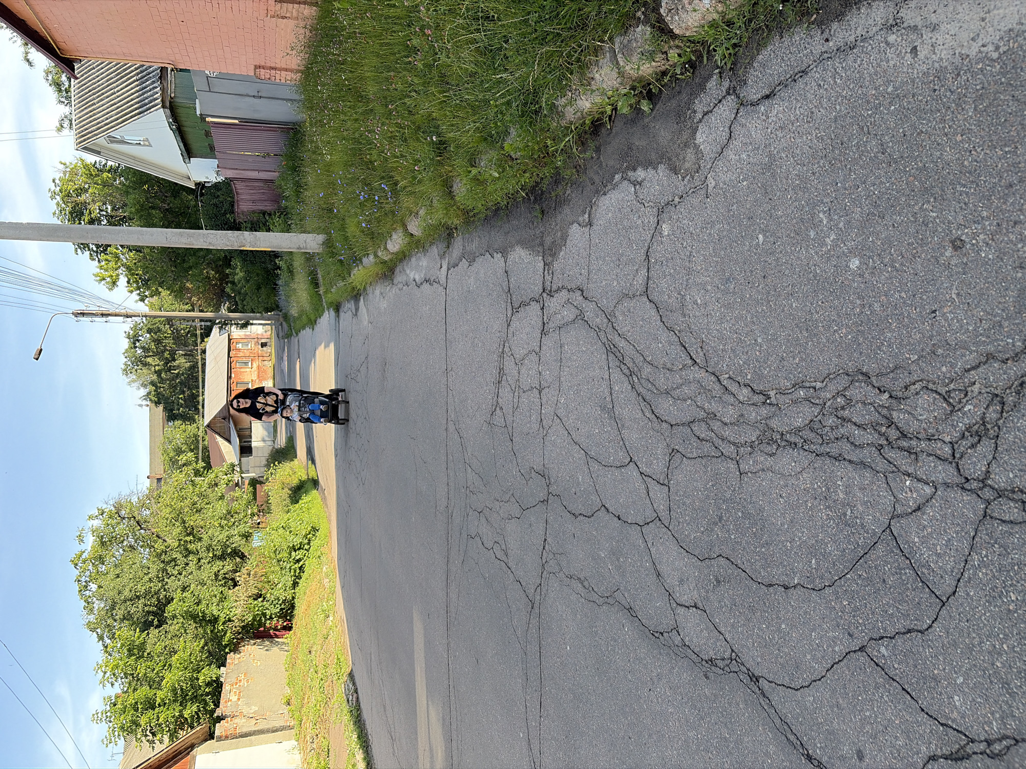 A woman is pushing a stroller down a cracked asphalt road in Chernihiv, Ukraine. The woman is wearing a black t-shirt and dark pants, and her hair is dark and pulled back. A child, wearing a blue shirt and gray pants, is in the stroller. The road is lined with houses, trees, and grass. The sky is blue with some clouds. The sun is shining, casting shadows on the road.