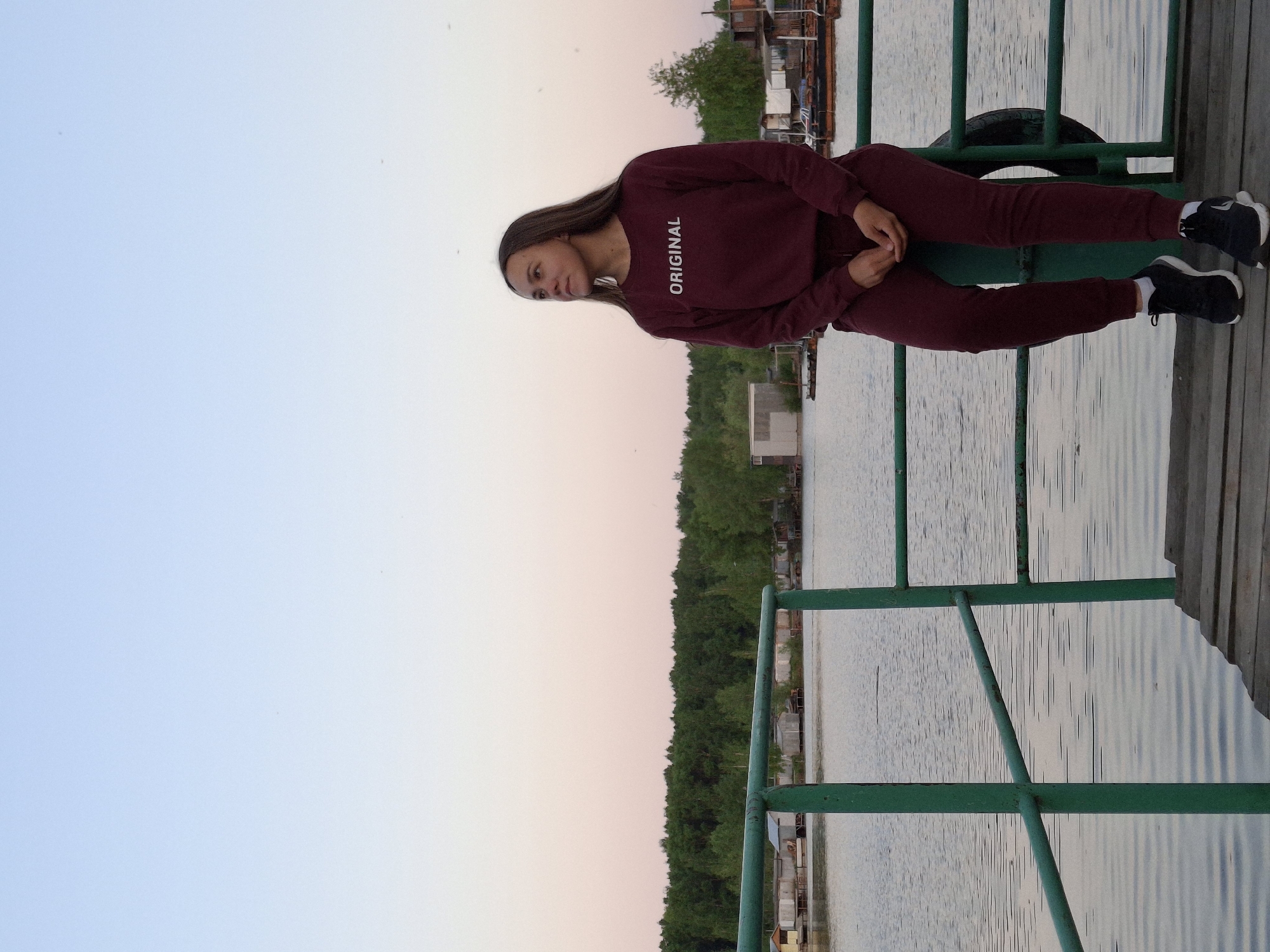 A woman is sitting on a green railing on a wooden deck overlooking a body of water in Chaikovsky, Russia. The woman is wearing a maroon sweatshirt with the word "ORIGINAL" printed on it, maroon pants, and black sneakers with white socks. She is looking to her left. In the background is the water, a treeline, and buildings. The sky is a gradient of blue and pink.