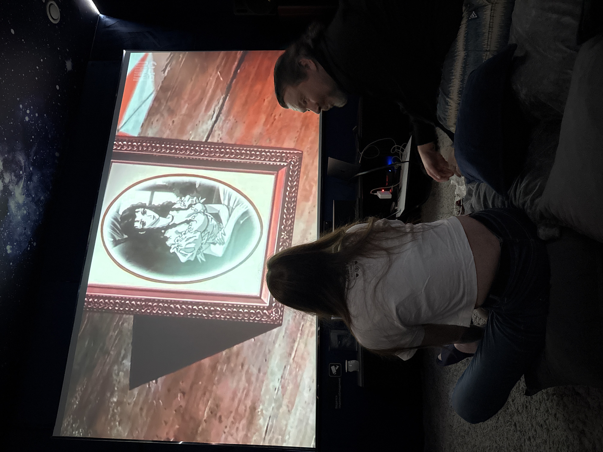 Here's a forensic description of the image:
**Scene:** The image depicts a room with a large screen displaying an image of a framed portrait. A man and a woman are partially visible in the foreground, seemingly looking at the screen.
**People:**
* **Man:** The man is standing to the right of the screen. He appears to be wearing a dark-colored shirt or jacket.
* **Woman:** The woman is located at the bottom center of the image. She is wearing a white t-shirt and jeans.
**Subjects:**
* **Screen:** A large screen displaying an image of a framed portrait. The portrait seems to be a black-and-white image of a woman in a Victorian-era style.
* **Wall:** To the left of the screen, the wall is painted with a starry galaxy theme.
**Objects:**
* **Cables and Electronics:** A cluster of cables, electronics, and adapters are visible near the screen, likely related to the screen or projection setup.
* **Couch:** A couch or seating area is positioned to the right of the screen.
* **Pillows:** At least two pillows are on the couch.
* **Picture:** The framed picture depicts an oval portrait of a woman in what appears to be Victorian-era attire.
**Actions and Interactions:**
* The man and woman are both oriented toward the screen, suggesting they are watching or observing something displayed on it.
**Environment:**
* The lighting in the room is subdued. The room appears to be a living room or entertainment area.
**Location:**
* There is no information in the image to indicate it is in Moscow, Russia.