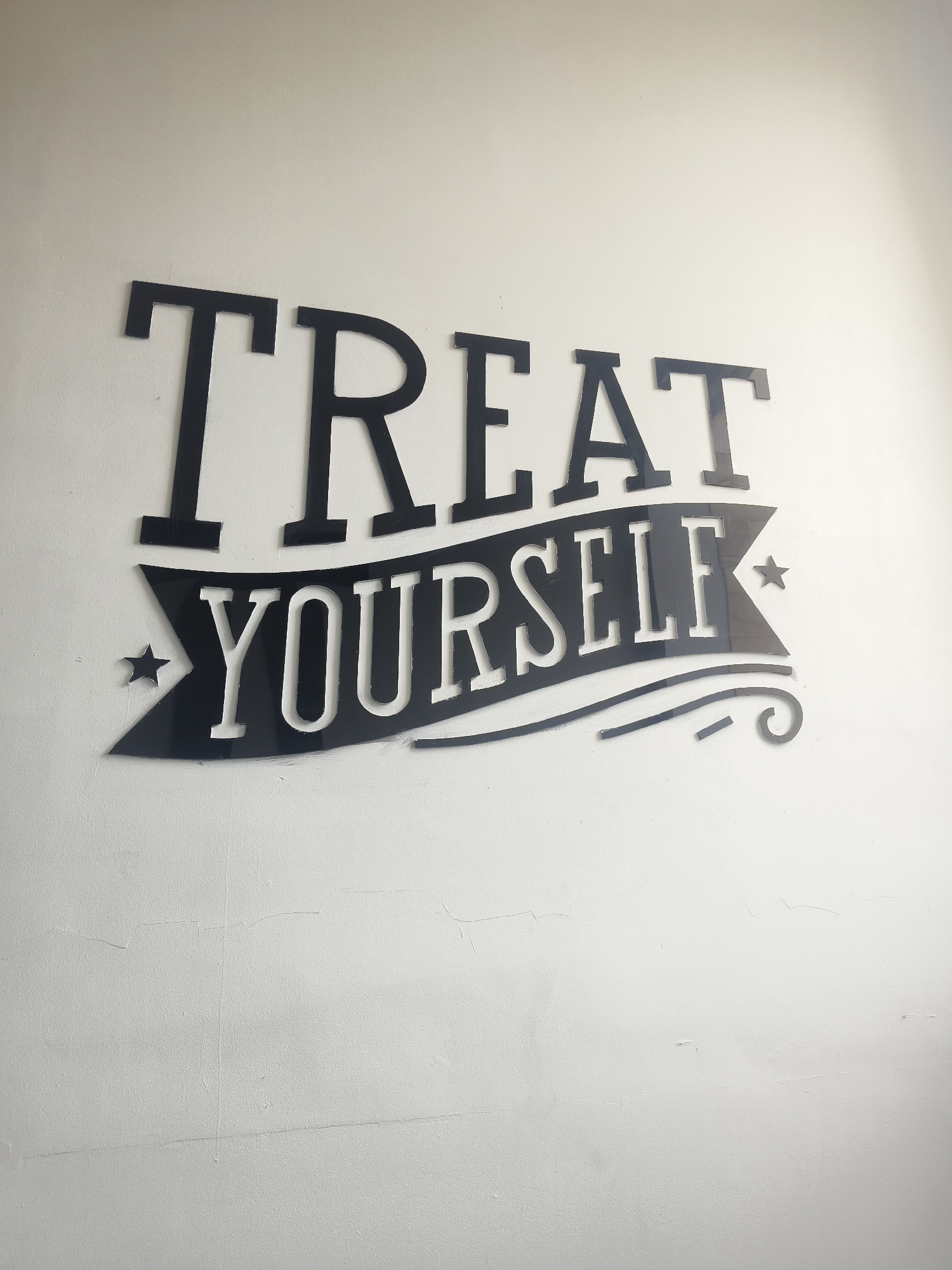 A black, cut-out acrylic sign with the words "TREAT YOURSELF" is mounted on a textured, off-white wall. The sign is in a decorative font, with "TREAT" in large capital letters above a banner that contains "YOURSELF." The banner is adorned with small star shapes and swirling decorative lines. The wall appears to be an interior surface, possibly in a commercial establishment, given the nature of the sign. There are some subtle imperfections and cracks on the wall's surface. The lighting suggests an indoor environment with light coming from an unseen source above and to the right. The image is captured from a slightly upward angle, focusing on the sign and the wall.
