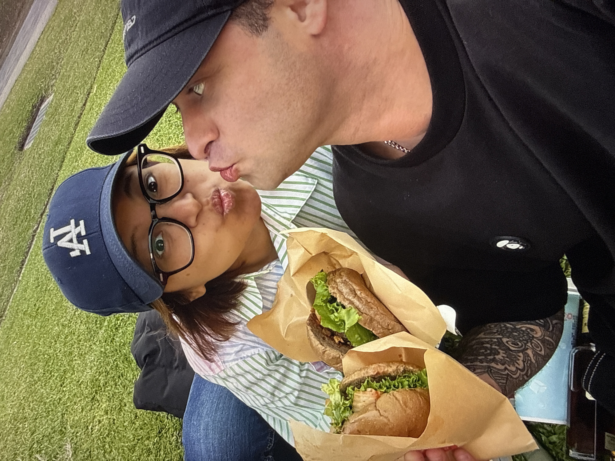 The image shows a young couple enjoying a meal on a grassy area. The woman, wearing a blue baseball cap with a white "LA" logo and glasses, is holding two sandwiches wrapped in brown paper. She is puckering her lips as if to kiss. The man, wearing a black baseball cap and a black t-shirt, is leaning in towards her, also puckering his lips. He has a tattoo on his left arm. The background appears to be a park with artificial turf. The lighting suggests it is daytime. There is no visible indication of the specific location within Taipei, Taiwan.