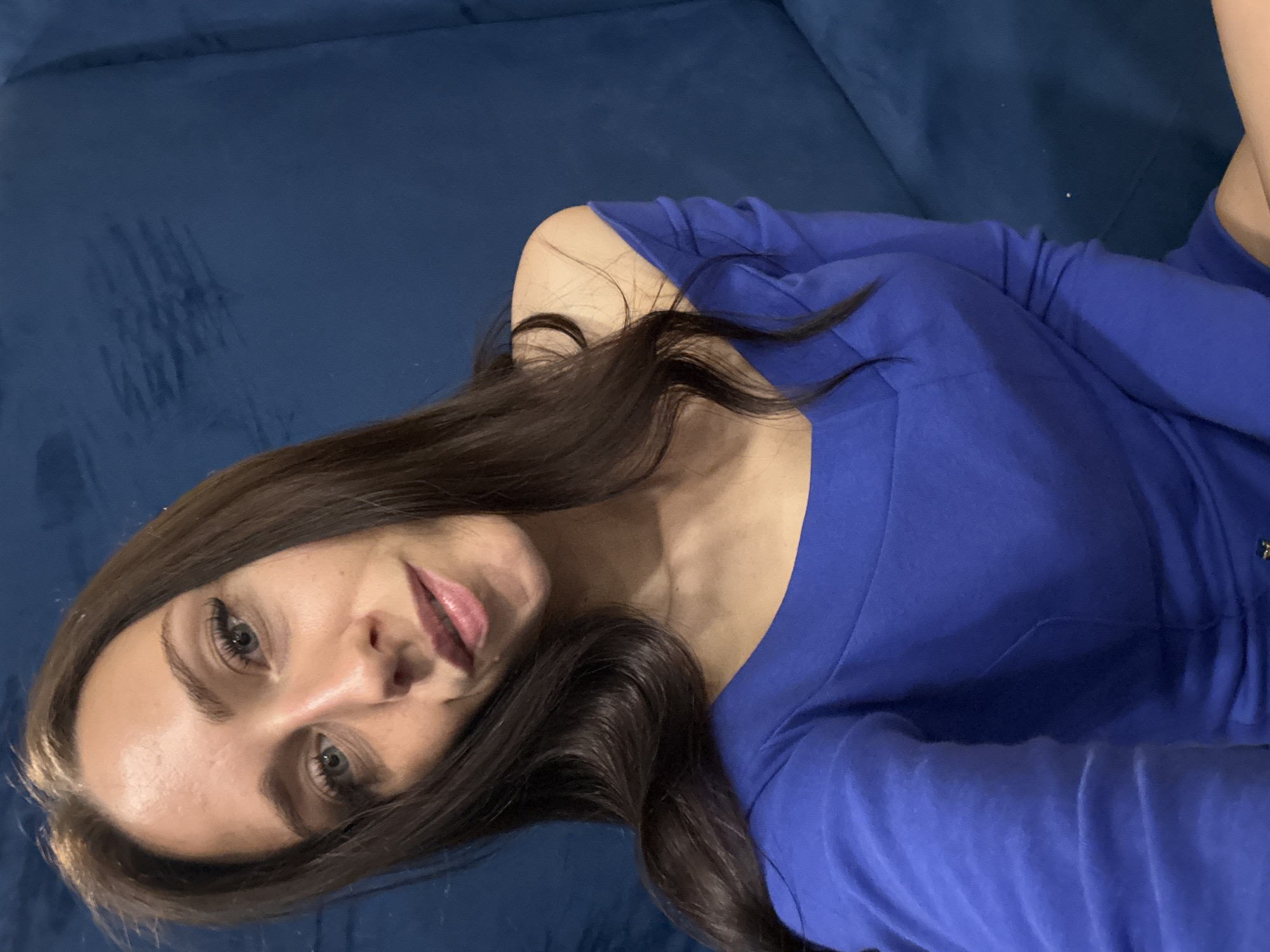 A young woman with long, dark brown hair and fair skin is captured in a selfie. She has striking blue eyes and is wearing a vibrant blue off-the-shoulder top. The background is a textured, dark blue surface, possibly a couch or upholstered wall. The lighting is soft, highlighting her features and creating a gentle glow. The setting appears to be indoors, possibly a home or studio. There is no indication of the specific location within Saint Petersburg, Russia.