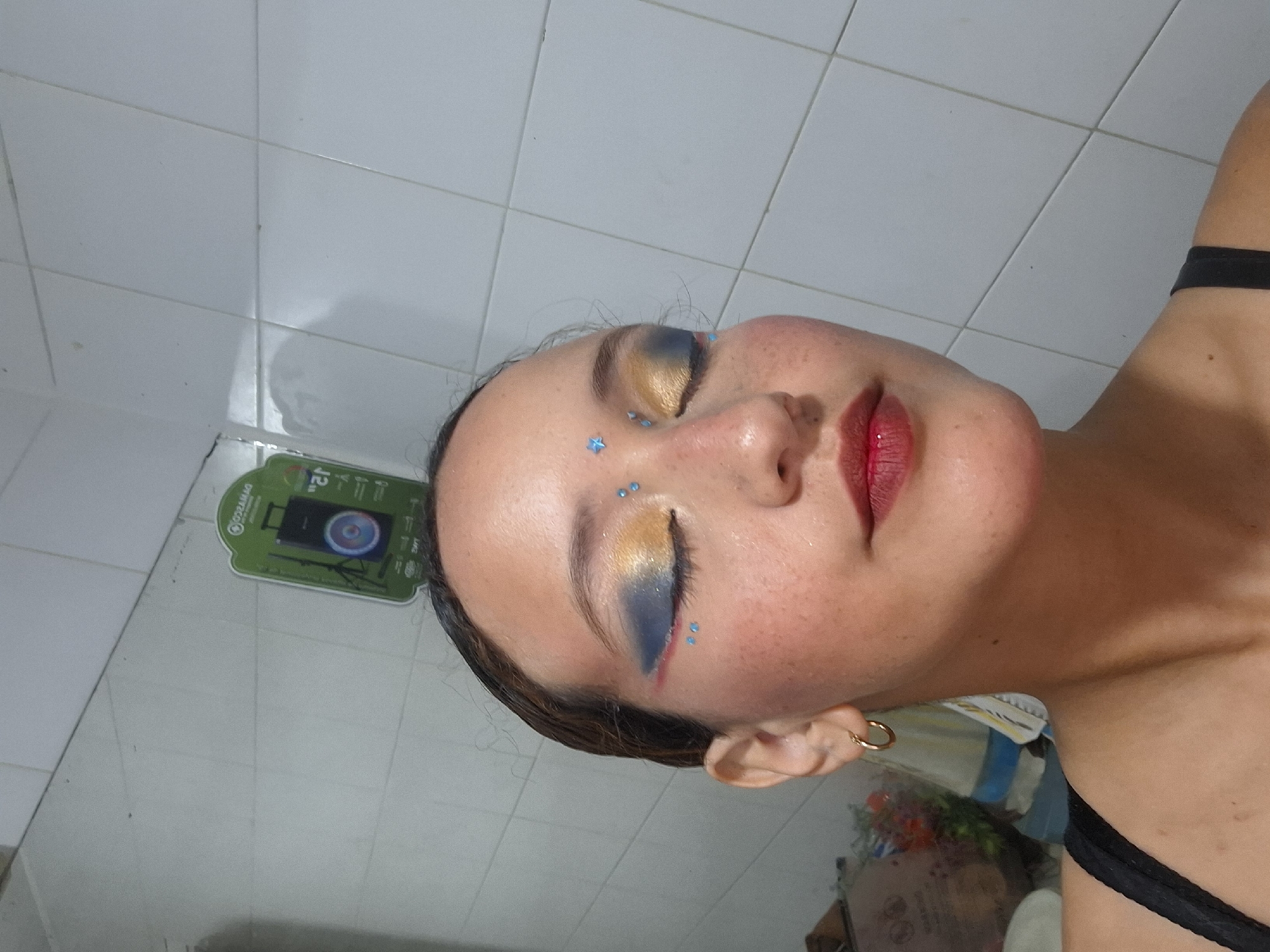 A selfie-style photograph shows a person with fair skin, presumably female, with unique makeup. The eyes are adorned with a blue and gold color scheme, accented with small blue star-shaped details on the brow and cheek. The person's lips are painted in a vibrant red. A simple gold hoop earring is visible. The person is wearing a black camisole or tank top. The background appears to be a tiled bathroom. The location is not discernible from the image provided. It is not possible to tell the location is in Playa El Agua, Venezuela.