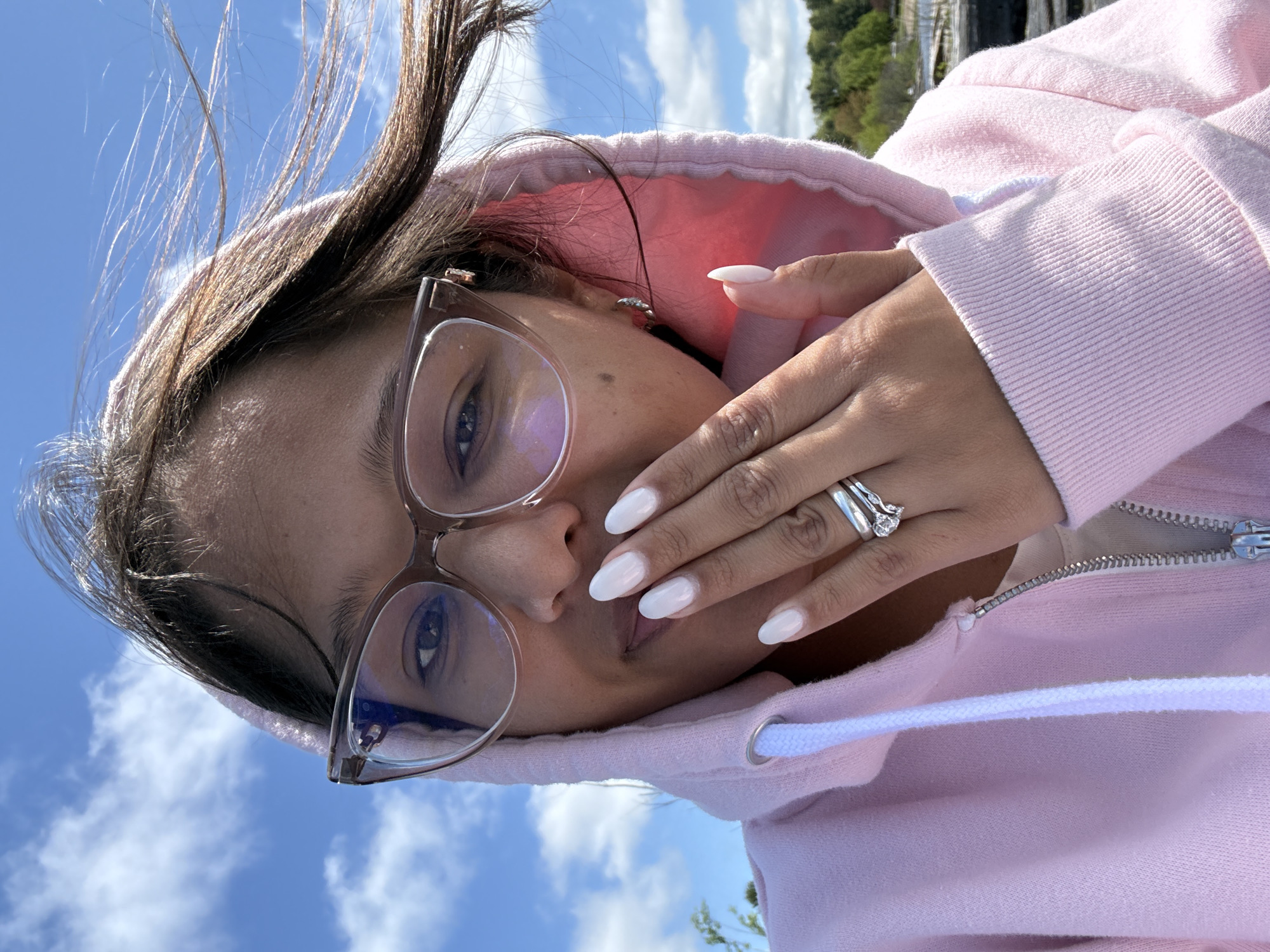 A woman with brown hair and light skin is seen in a close-up, angled shot. She is wearing eyeglasses with transparent frames and a pink hooded sweatshirt. Her left hand, adorned with white nails and several rings, is positioned near her face. The background features a blue sky with white clouds. The location is Unknown, Unknown.