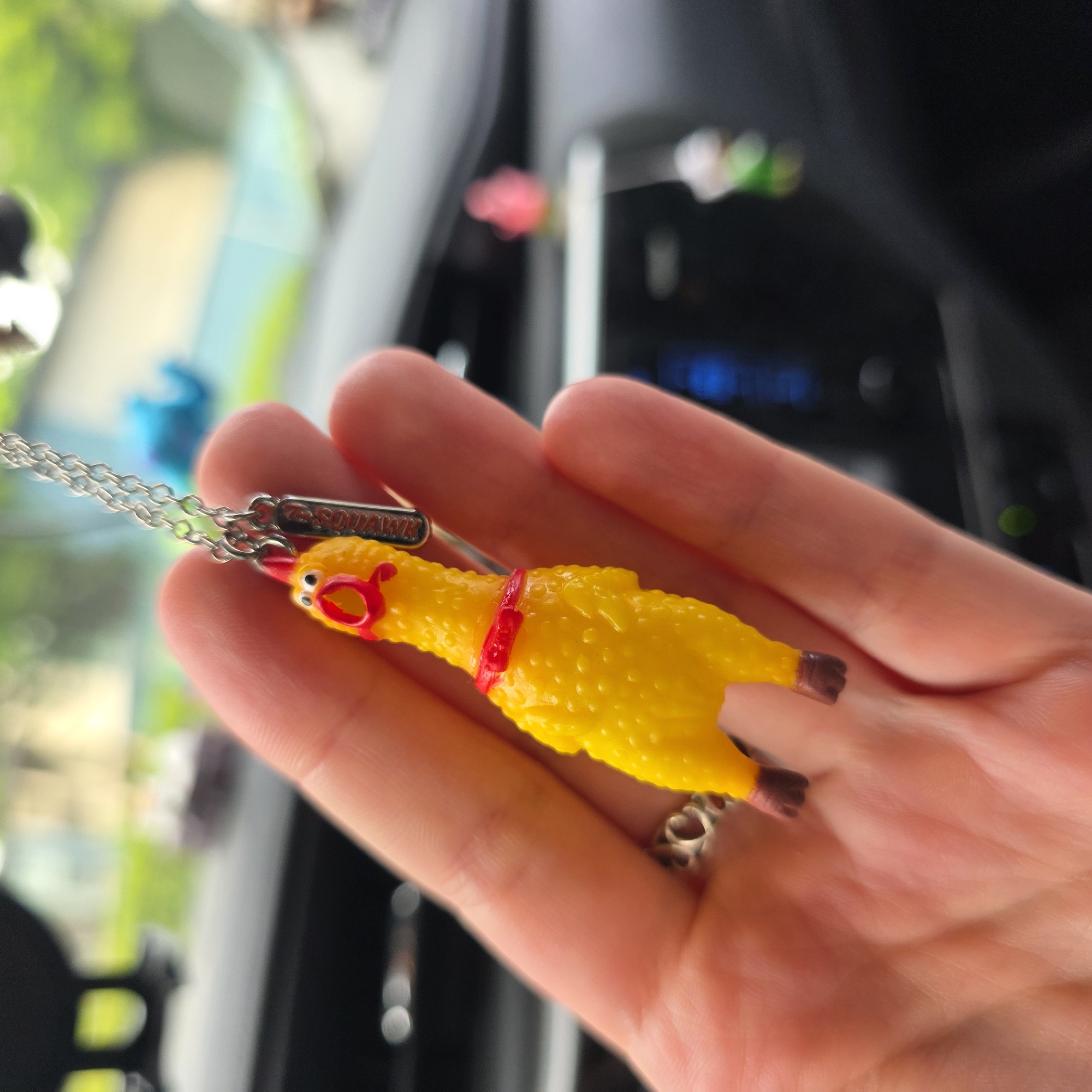 In Portland, Oregon, USA, a hand with a silver ring is holding a yellow rubber chicken keychain. The chicken has a red collar and a red comb, with a tag hanging from its neck. The background shows the interior of a car, with the dashboard and a blurred view of the outdoors through the windshield.
