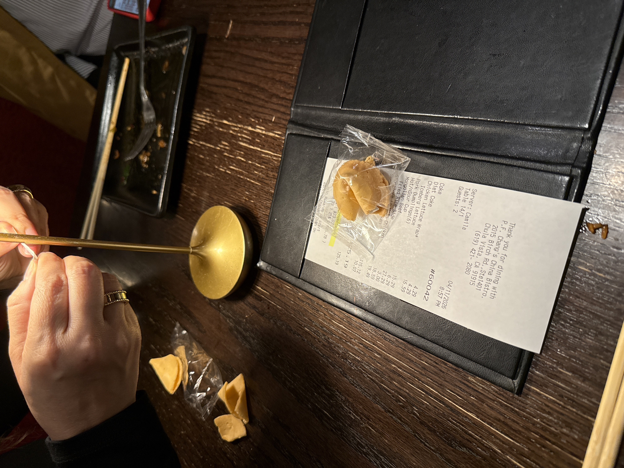 A restaurant scene in Chula Vista, California, United States of America shows a table with a black bill holder on a wooden surface. The bill holder contains a bill from P.F. Chang's China Bistro at 2015 Birch Rd., Ste 1401, Chula Vista, CA 91915, showing a date of 04/11/2026 and a check number #60042. Next to the bill is a sealed fortune cookie in a plastic bag. A person with a gold ring on their finger is holding a chopstick. Next to their hand are open fortune cookies in transparent plastic. There is also a nearly finished plate of food with chopsticks beside it.