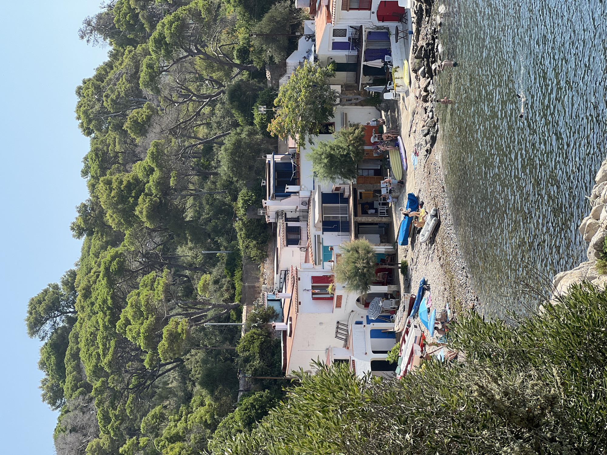 A high-angle medium shot captures a coastal scene in Palamós, Spain, featuring a collection of whitewashed houses with terracotta roofs nestled against a densely forested hillside. The foreground is dominated by a small, pebbly beach dotted with various boats, including several small dinghies, a red and white vessel, and a blue inflatable. Some of these boats are resting on the shore, while others are partially submerged in the clear, rippling water. Several people are visible on the beach and in the water. On the beach, a group of individuals, some in swimwear, are engaged in leisure activities, with one person lying under a blue umbrella, and others interacting near a yellow inflatable kayak. In the water, two individuals are swimming, one with snorkeling gear. The houses are characterized by their simple architecture, with colorful shutters on some windows and balconies. Lush green trees and foliage cover the hillside that rises behind the houses. The sky is a clear, pale blue.