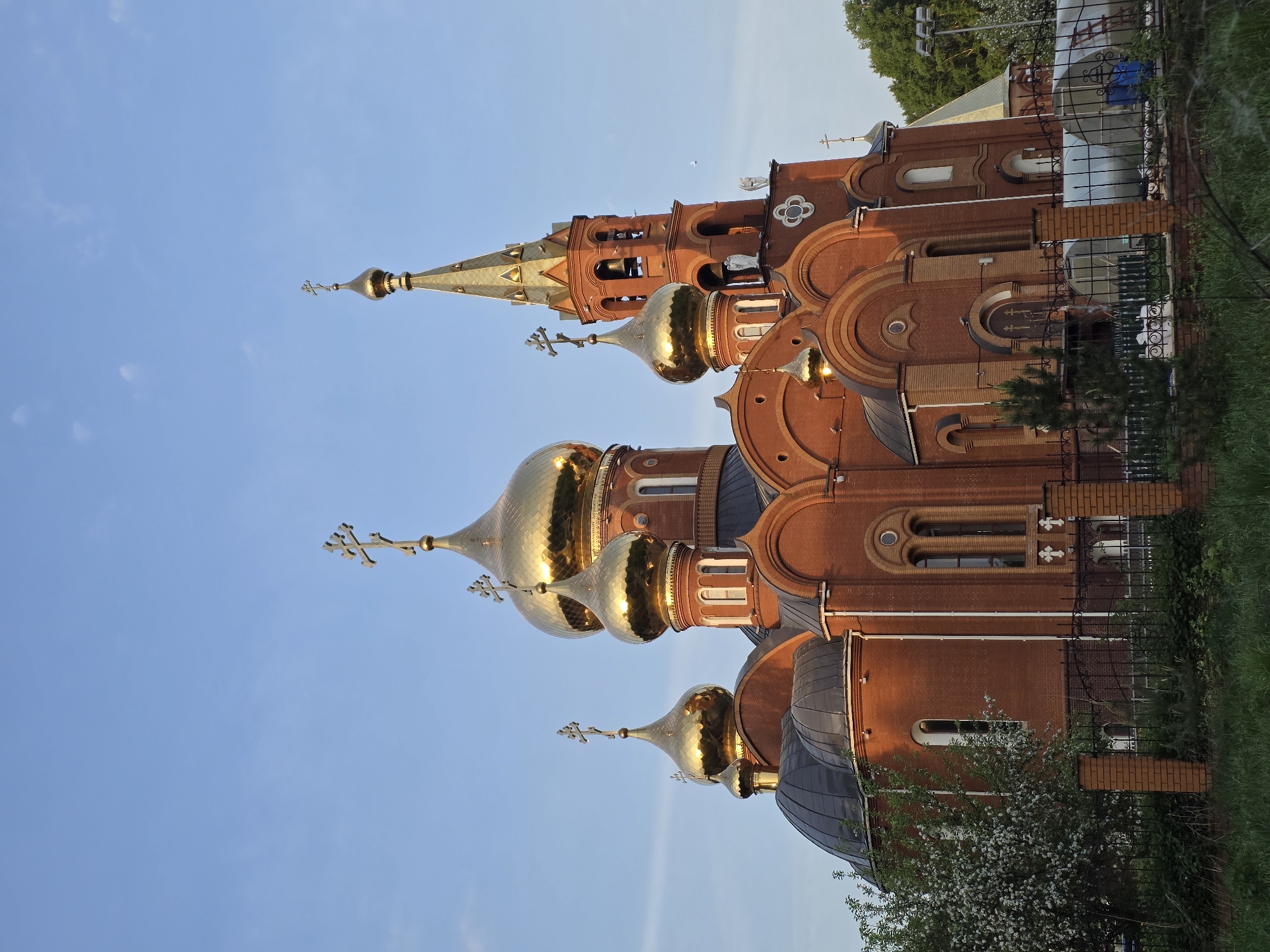 The image shows an exterior view of the Church of St. George the Victorious, located in Chaikovsky, Russia. The church is constructed of red brick and features a distinctive architectural style with multiple golden onion domes topped with crosses. The domes are reflecting sunlight. The sky is clear and blue. In the foreground, there is vegetation including grass and flowering trees. A wrought iron fence separates the church grounds from the surrounding area.