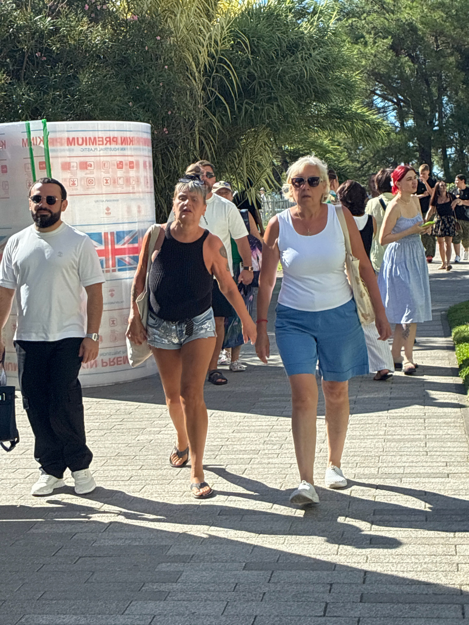 In Sochi, Russia, on a sunny day, a group of people walk along a paved pathway. In the foreground, two women walk side-by-side. The woman on the left, with short blonde hair and sunglasses, wears a white tank top and blue denim shorts. She carries a light-colored tote bag over her shoulder. The woman on the right, with short dark hair and sunglasses, wears a black tank top and denim shorts with sequin detailing. She also carries a tote bag. To the left of the women, a man with dark hair and a beard, wearing a white t-shirt and black pants, walks ahead. He wears sunglasses and a watch. Behind them, other people are visible walking in the same direction. To the left of the man, a large white cylindrical object with text and images printed on it is partially visible. The background features lush green trees and foliage. The ground is paved with gray rectangular tiles, and the sunlight casts long shadows.