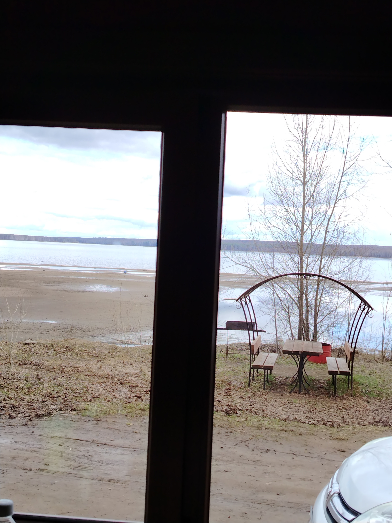 From an indoor perspective through a window frame, the scene unfolds as follows:
The location is most likely in the city of Chaikovsky, Russia based on the surroundings. The outside features a sandy bank leading to a large body of water, possibly a lake or wide river, stretching into the distance under a cloudy sky. The opposite bank is lined with trees, indicating a forested area. Closer to the window, there is a patch of land covered with dry leaves. There is a wrought-iron archway, under which are a wooden table and benches, and a grill. A white car is partially visible in the bottom right corner. The indoor perspective is from a dimly lit area.