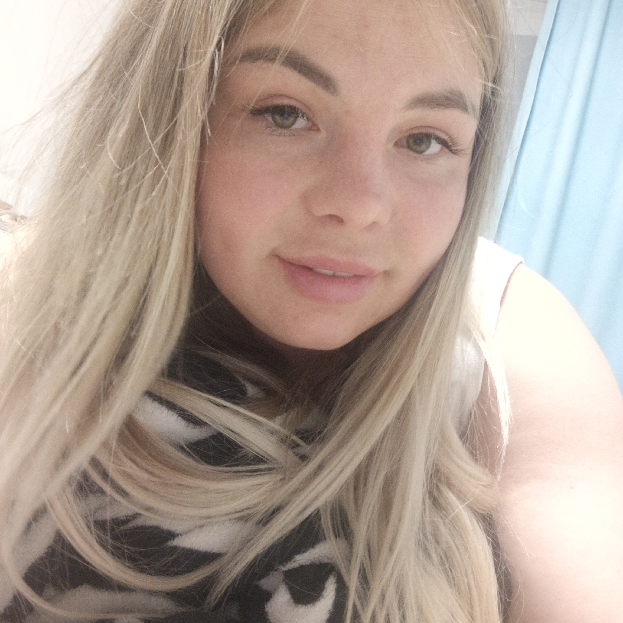A close-up selfie depicts a young woman with light blonde hair and fair skin. She has a slightly off-center pose, gazing directly at the camera. Her hair is tousled, with strands falling across her face and shoulders. She has light-colored eyes and natural-looking eyebrows and eyelashes. The woman has a subtle smile and slightly parted lips. She is wearing a white tank top. In the foreground, a black and white patterned garment is partially visible. In the background, a light blue curtain hangs vertically. The location is unknown.