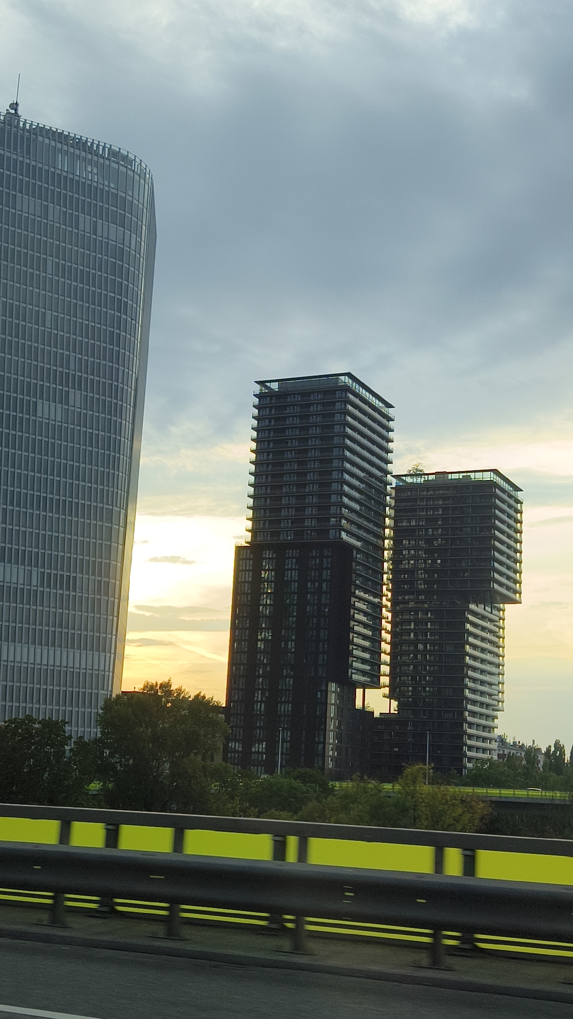 The image shows a modern cityscape dominated by several tall buildings. On the left, a large, curved skyscraper with a grey, grid-like facade stands prominently. To the right of this, two darker, rectangular skyscrapers are visible, one slightly taller than the other, with numerous balconies and glass windows. The sky is overcast with hints of sunlight breaking through. In the foreground, a highway railing with bright yellow accents runs horizontally across the lower part of the image. Trees and vegetation can be seen between the highway and the buildings, suggesting an urban park or green space. The scene appears to be captured from a moving vehicle on the highway. The buildings' architecture suggests a contemporary urban development. This scene is consistent with views of the Donau City district in Vienna, Austria, known for its modern skyscrapers.