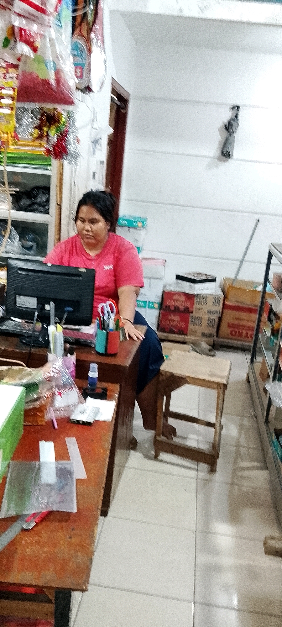 The image depicts a woman in a red t-shirt sitting at a desk in what appears to be a small shop or office. She is looking down at papers or a computer. On the desk, there is a computer monitor, a pen holder with pens and scissors, a bottle of blue liquid, and various papers and plastic bags. In the background, there are shelves filled with items, boxes stacked up, and a small wooden stool. The room has white walls and a tiled floor. A small decorative figure is mounted on the wall. The lighting is artificial, suggesting an indoor environment. The overall scene suggests a workspace where the woman is engaged in some administrative or retail task. The image does not provide enough visual information to determine the specific location within Satriamekar, Indonesia, beyond it being an indoor commercial establishment.
