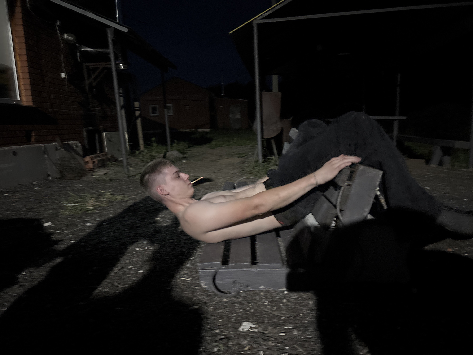 The image depicts a young man lying on a wooden bench outdoors at night. He is shirtless and wearing dark pants. His body is positioned as if he is performing an exercise, with his hands holding onto a grey object. A cigarette is visible in his mouth. The background shows a brick building to the left and other structures that suggest an outdoor recreational area or a rural setting. The lighting is dim, casting strong shadows. The scene appears to be in Chaikovsky, Russia.