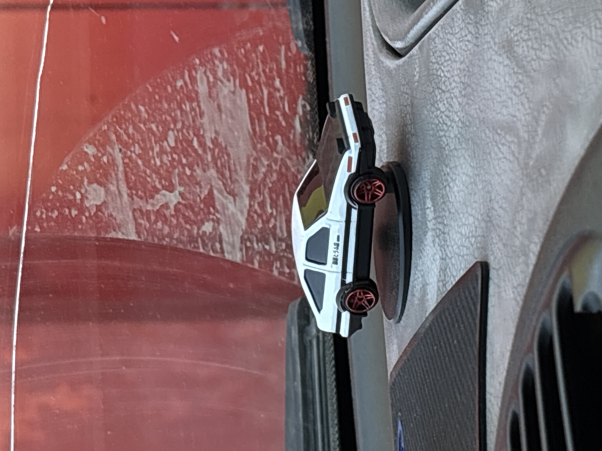 The image shows a white toy car with red wheels attached to a gray textured surface, possibly a car interior. To the left of the car is a dirty red panel, likely a car door or dashboard. Other parts of the vehicle interior are also visible. Location information is not present.