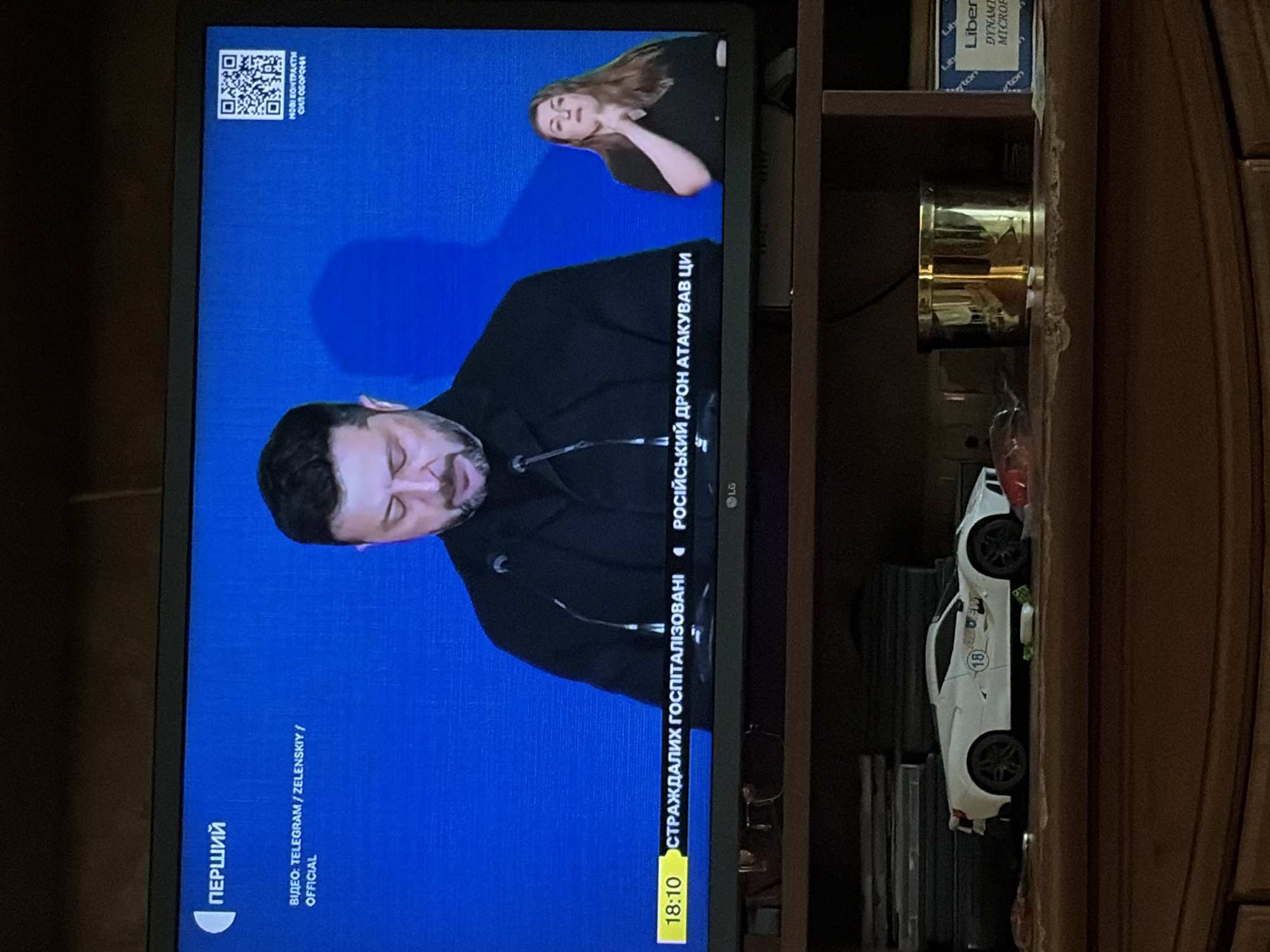 The image captures a television screen displaying a news broadcast. On the screen, a man, identified as President Zelenskyy, is speaking at a podium. To the right of the screen, a woman is visible, likely an interpreter providing sign language. The screen shows a blue background with text and a QR code in the upper right corner. A ticker tape at the bottom of the screen displays the time as "18:10" followed by Ukrainian text. The television is placed on a wooden shelf, and below the shelf, a white toy sports car is visible. There are also several other objects on the shelf, including a metallic container and a box with text on it. The overall environment appears to be an indoor setting.