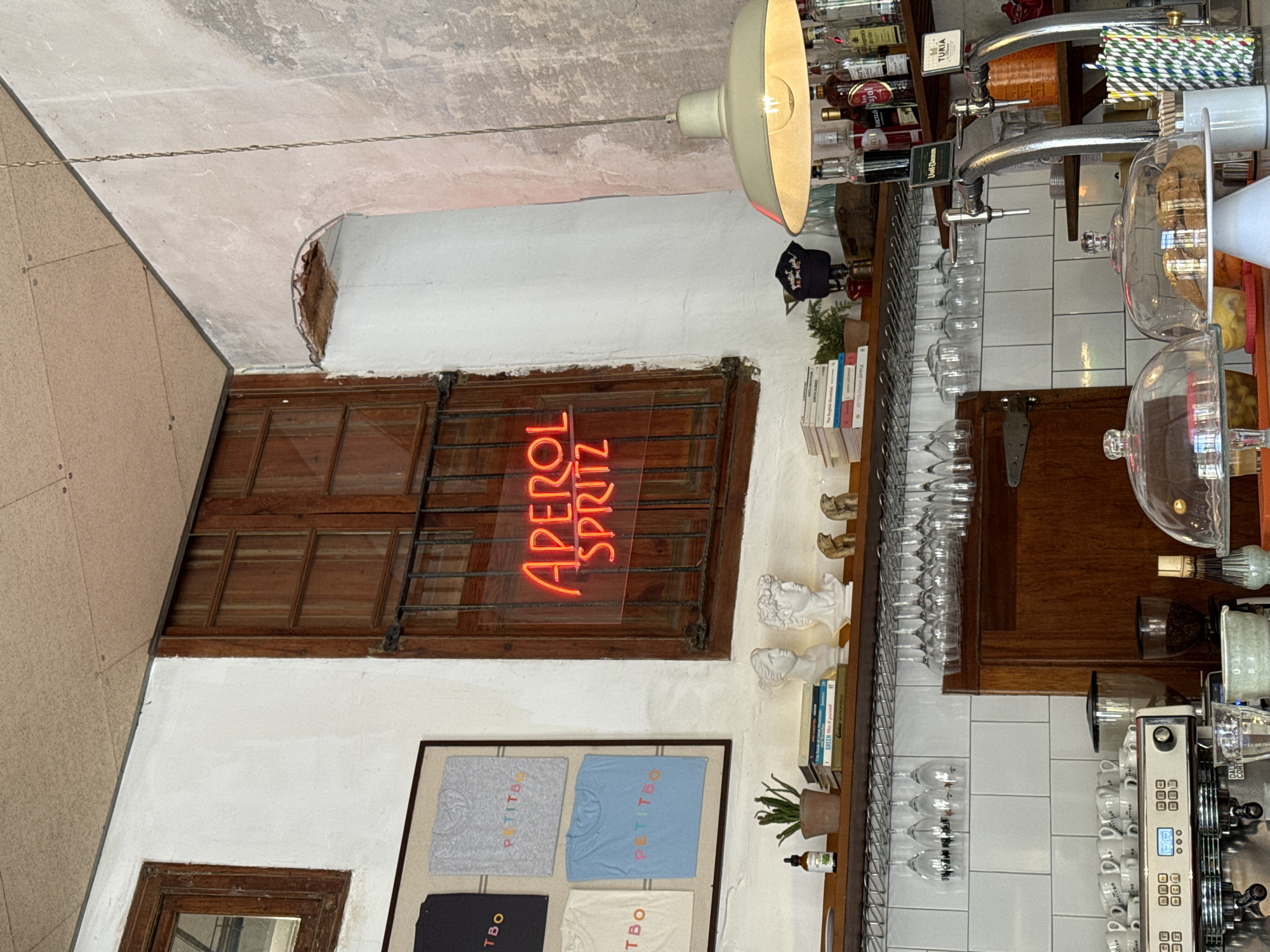 The image shows the interior of a bar or cafe, likely in Barcelona, Spain. A closed wooden window with metal bars and an "Aperol Spritz" neon sign is visible. Below the window is a framed display of "Petitbo" t-shirts. A shelf to the right of the window holds books, small sculptures, and plants. The bar area includes rows of wine glasses, liquor bottles, pastry displays, and a coffee machine. The style suggests a modern, European aesthetic.