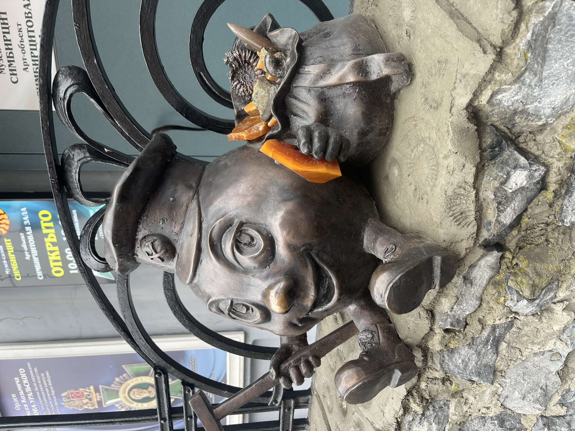 This image shows a bronze sculpture of a miner's head emerging from a rough, stone-like wall. The sculpture is mounted on a black, ornate iron gate. On top of the head is another bronze sculpture of a man holding a piece of amber.
The backdrop includes signage in Russian, which translates to "Sim bir Site" and mentions being an "Art Object" and the "Sim bir Site Hall." The sign also indicates that it is open from 10:00 AM.
It is a tourist attraction dedicated to Simbirsk - Simbirzit mineral. It's located on Lenin Street in Ulyanovsk, Russia.