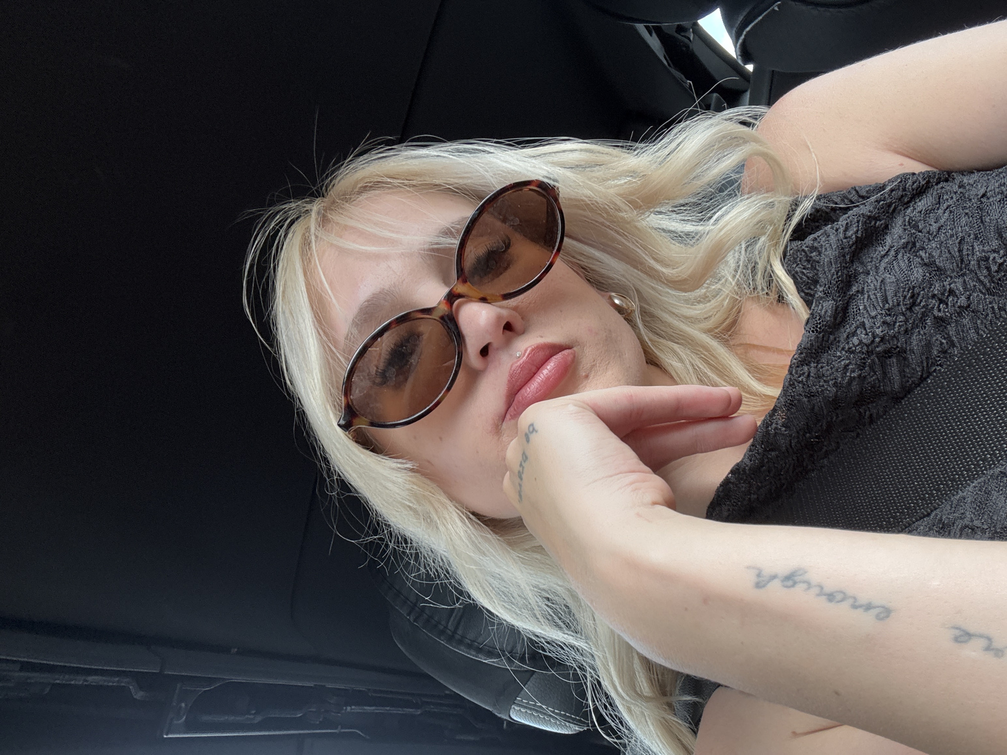 An overhead, close-up shot captures a young white woman with blonde hair and blue eyes looking down. She is wearing tortoiseshell sunglasses and a black lace top. Her left hand rests on her chin, and a tattoo is visible on her inner left forearm that says "be brave." The woman is seated in the passenger seat of a car, with the black leather interior and part of the black seatbelt visible. The background is dark, suggesting it is inside a vehicle. The lighting is natural, coming from the front and side, creating highlights on her hair and skin. The woman's lips are slightly parted, and she has a small nose ring.