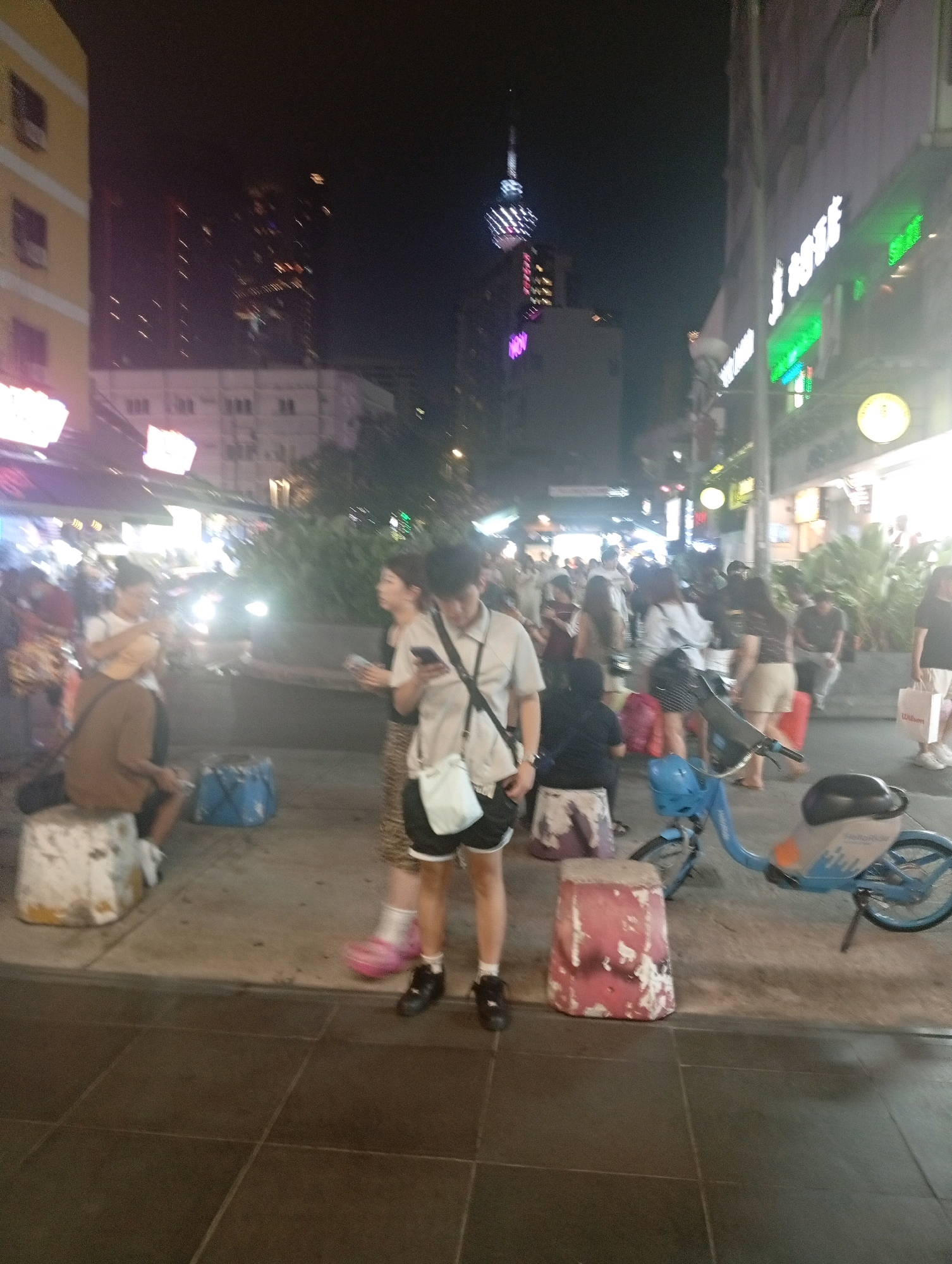 In this night-time scene in Kuala Lumpur, Malaysia, an outdoor market or street scene is depicted. In the foreground, a man in a light-colored short-sleeved shirt and dark shorts stands on a sidewalk. He is wearing black shoes and has a white bag slung over his shoulder. To his right, a woman in a patterned skirt and a light-colored shirt is also visible. She is holding a phone and appears to be interacting with the man. In the background, a blue bike-sharing bicycle is parked on the sidewalk. Several people are scattered throughout the scene, some walking and others sitting. The environment features buildings with illuminated windows and signs, suggesting commercial establishments. A prominent tower with lights is visible in the distance, likely the Kuala Lumpur Tower. The overall impression is that of a bustling urban environment at night.